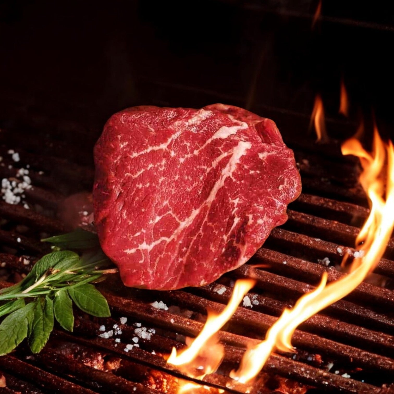 A 6oz Mino Prime Steaks filet sizzles on a hot grill, flames visible, adorned with fresh herbs and coarse salt scattered across the prime-grade beef.