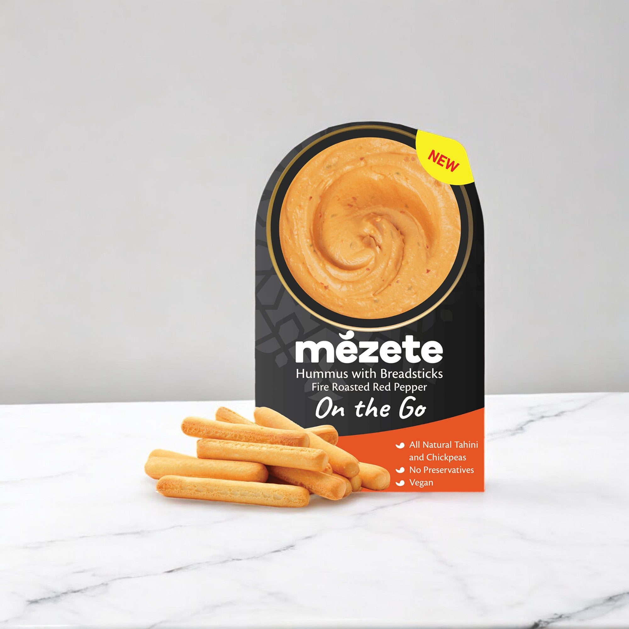 The packaging of Mezete's Fire Roasted Red Pepper Hummus with Breadsticks On The Go (3.2 oz - 1 Count) is highlighted, showcasing the container made with natural ingredients alongside several breadsticks on a marble surface.