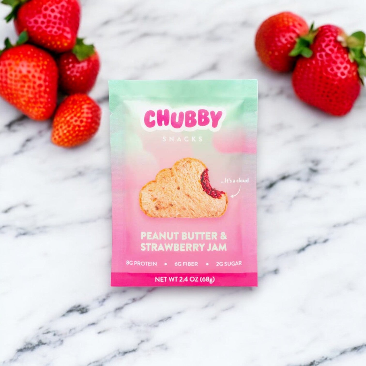 A packet of Chubby Peanut Butter & Strawberry Jam sits on a marble surface, surrounded by fresh strawberries. The packet highlights 8g protein, 6g fiber, and 2g sugar. Net weight is 2 oz.