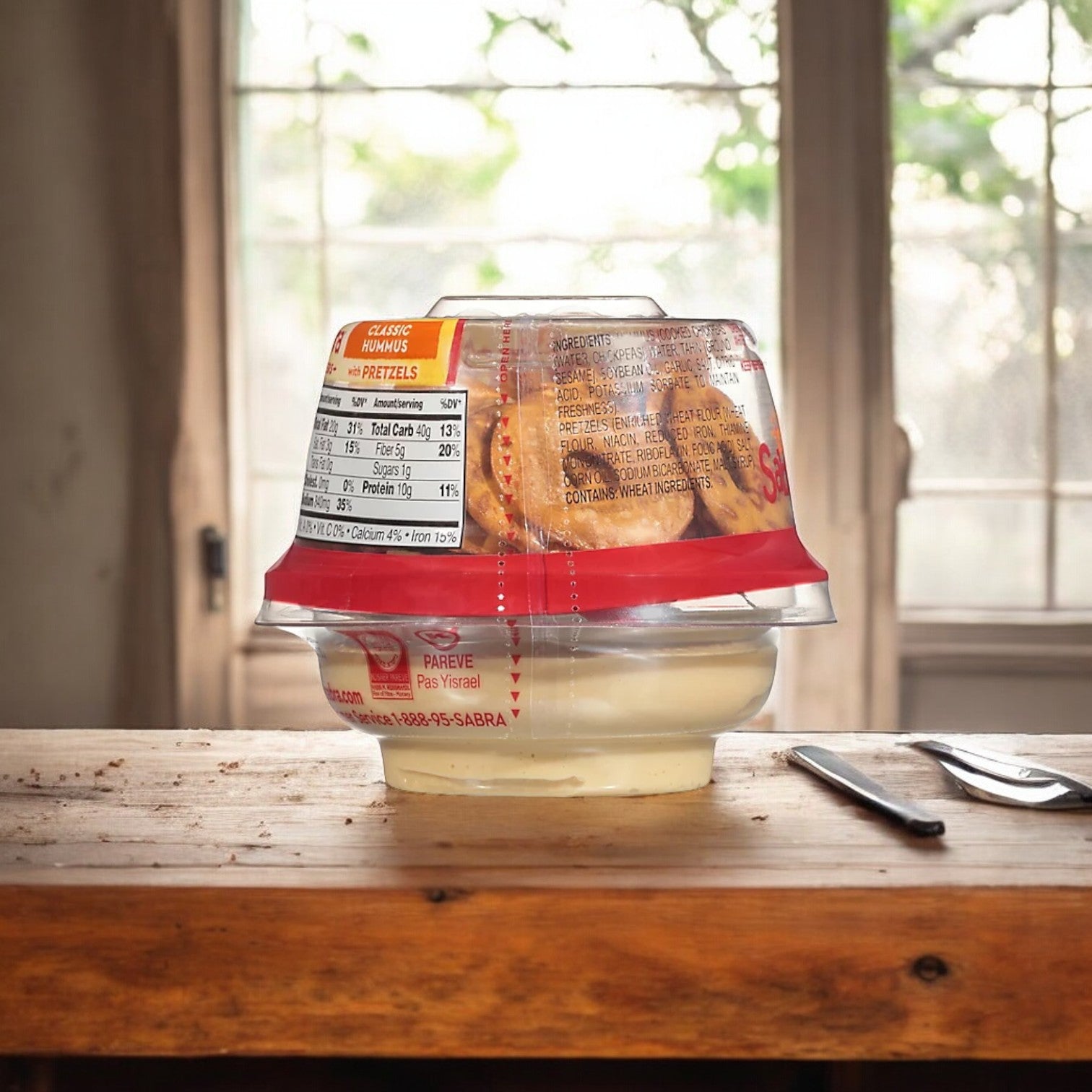 A container of Sabra Classic Hummus from the brand Sabra is placed on a wooden surface in front of a window. A spoon is lying on the table next to the creamy hummus dip with pretzels.