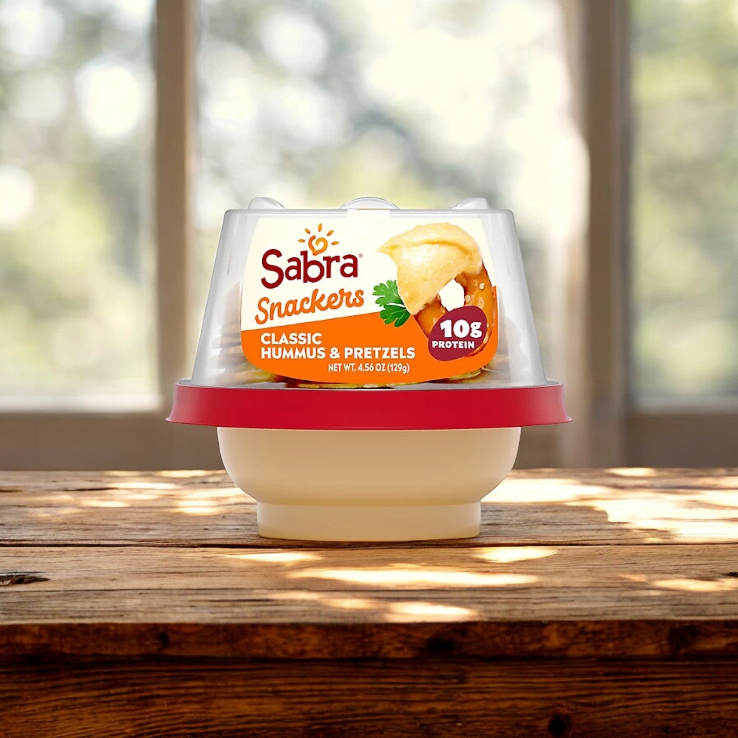 A package of Sabra Classic Hummus and pretzels from the Sabra brand sits on a wooden table with a blurred window view in the background, showcasing this versatile dip perfect for any occasion.