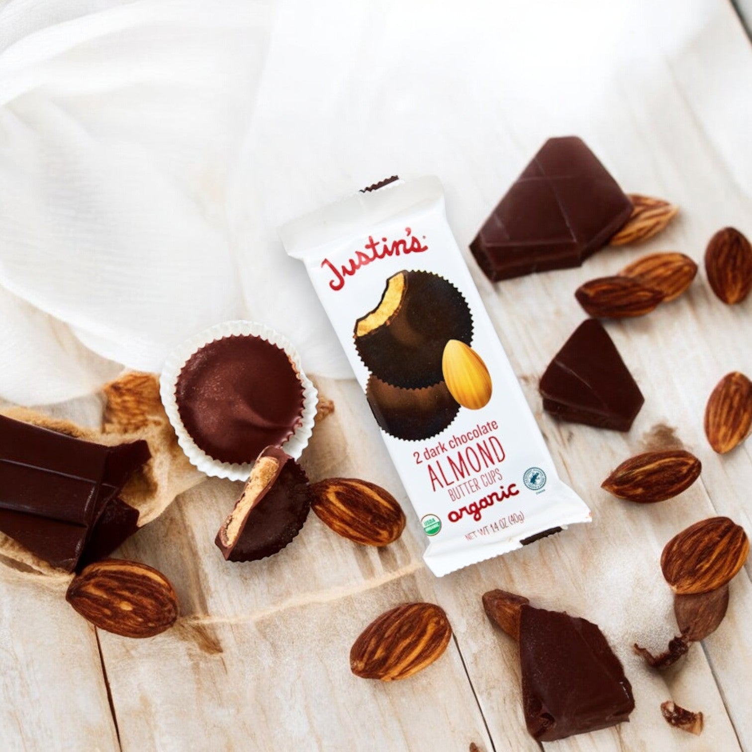 A package of Justin's Organic Dark Chocolate Almond Butter Cups-1.4 oz- 1 Count is placed on a wooden surface, surrounded by almonds, chocolate pieces, and some unwrapped almond butter cups from Justin's.