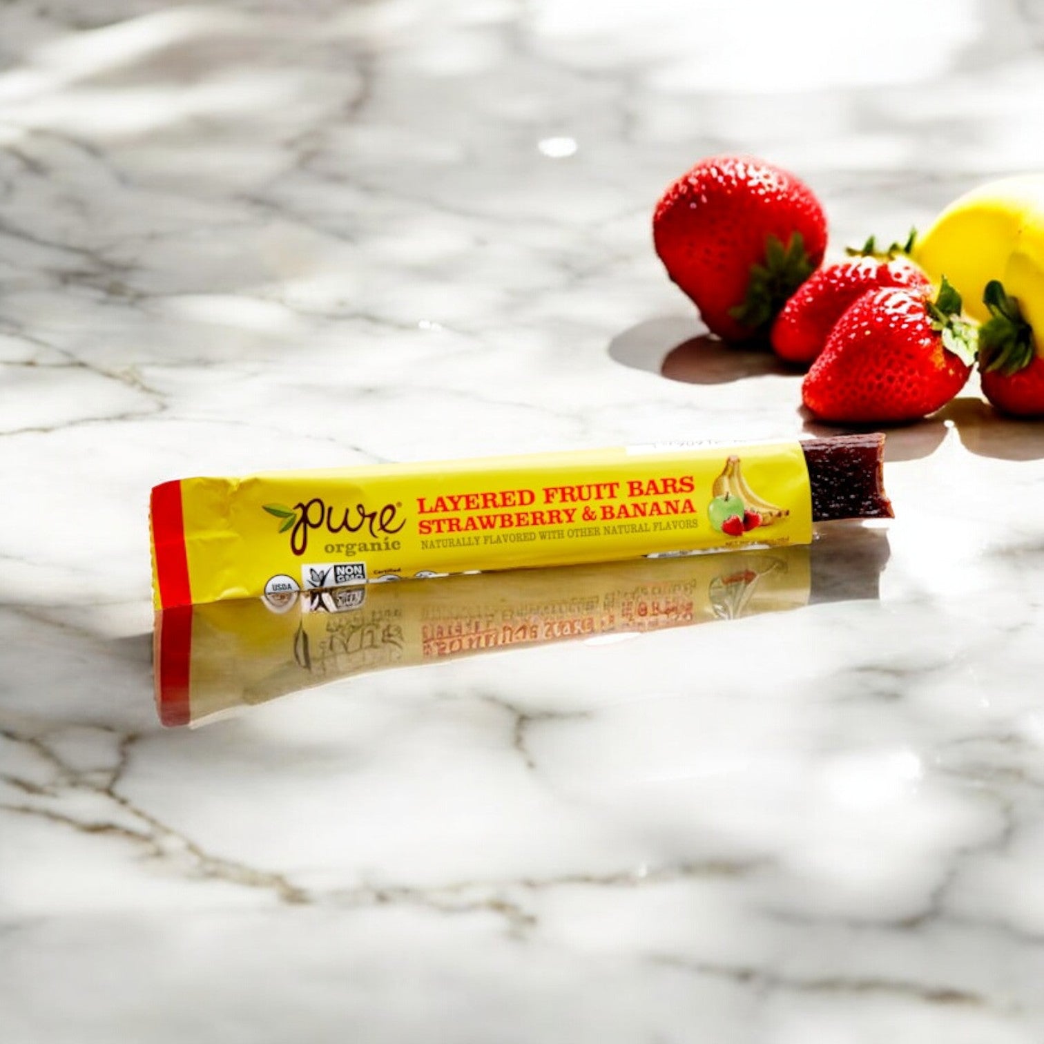 A Pure Organic Strawberry Banana Layered Fruit Bar, a vegan gluten-free snack from Pure Organic, is placed on a marble surface next to four fresh strawberries and a partially visible yellow fruit.