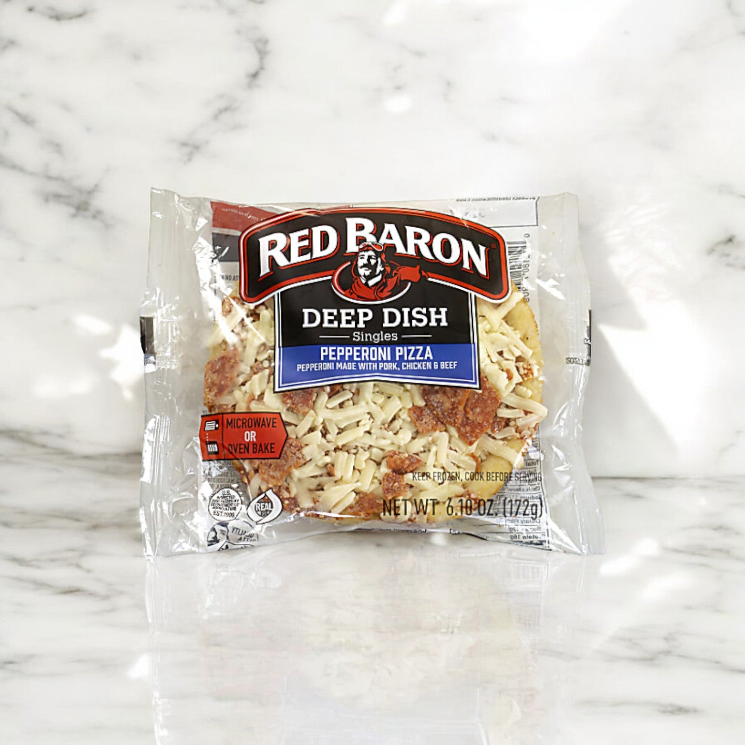 A packaged Red Barron Frozen Pizza Deep Dish Singles - 12 Count sits on a marble surface. The packaging highlights microwave and oven bake options, showcasing it as a quick and satisfying meal.