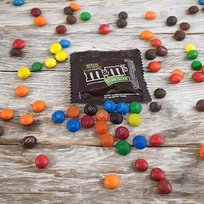 A variety of M&M candies scattered on a wooden surface, with an unopened pack of Mars Fun Size Chocolate M&M's - 5 Count Fun Size Packs at the center.