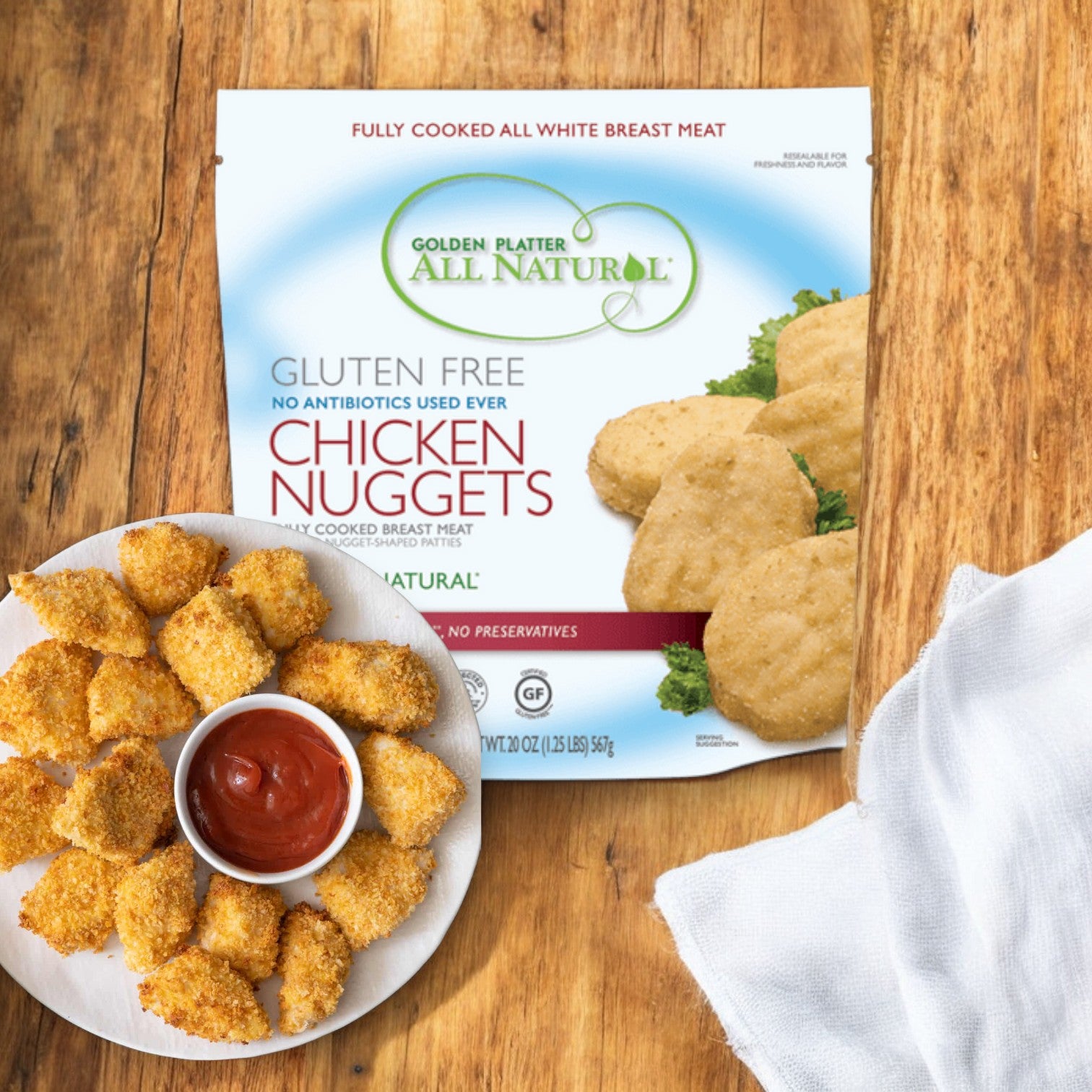 A package of Golden Platter Gluten Free Nuggets, 4 lbs, alongside a plate of cooked chicken breast nuggets with a red sauce for dipping on a white cloth.