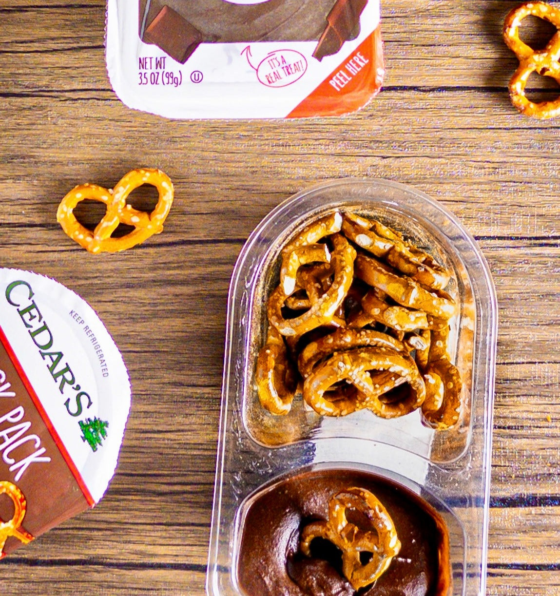 A Cedar's Snack Pretzels with Chocolate Hommus -1 Count tray sits on a wooden table, accompanied by another partially visible snack pack and scattered loose pretzels.