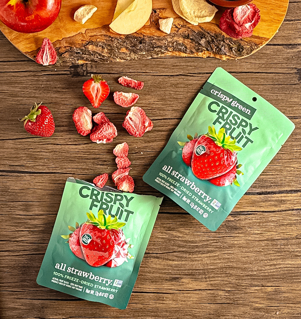 Two pouches of Crispy Fruit Freeze-Dry Strawberry - 0.35 oz - 1 Count are placed on a wooden surface near fresh and freeze-dried strawberries and apple slices, showcasing their natural sweetness for a guilt-free indulgence.