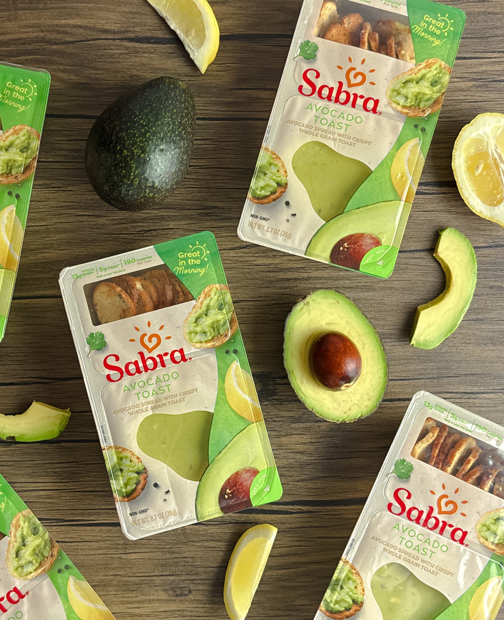 Several packages of Sabra Breakfast Avocado Toast are arranged on a wooden surface alongside fresh Hass avocado slices, lemon wedges, and a whole avocado, making for a perfect nutritious breakfast.