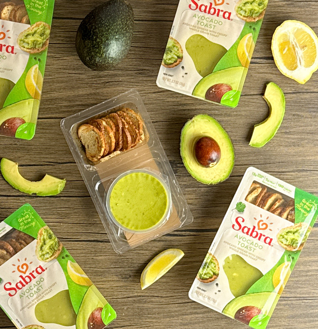 Image of Sabra Breakfast Avocado Toast packages, some open with toast slices, surrounded by whole and sliced Hass avocados, lemon slices on a wooden surface—perfect for a nutritious breakfast from the Sabra brand.