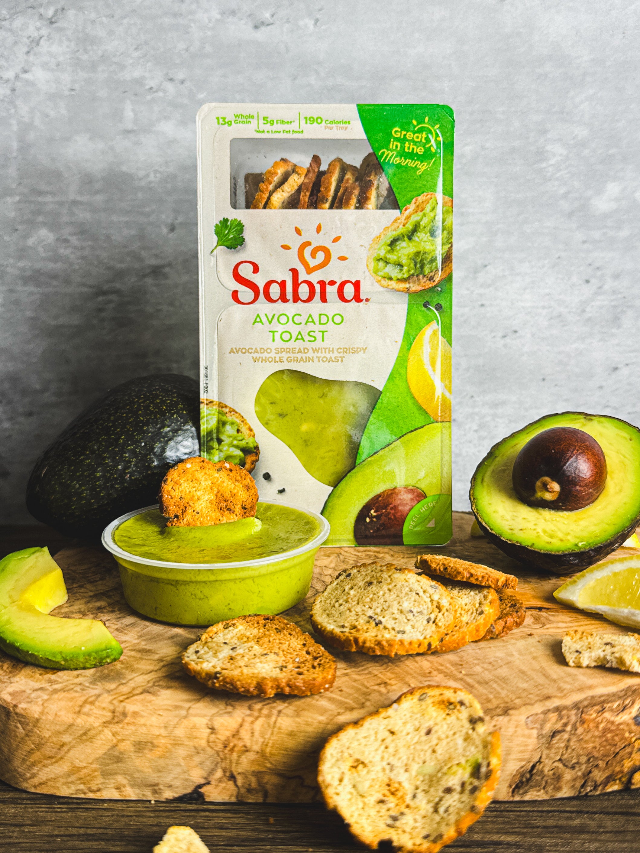 A nutritious breakfast, the Sabra Breakfast Avocado Toast package by Sabra includes a container of rich avocado spread, fresh Hass avocado slices, and hearty bread pieces on a wooden board beside whole and halved avocados.