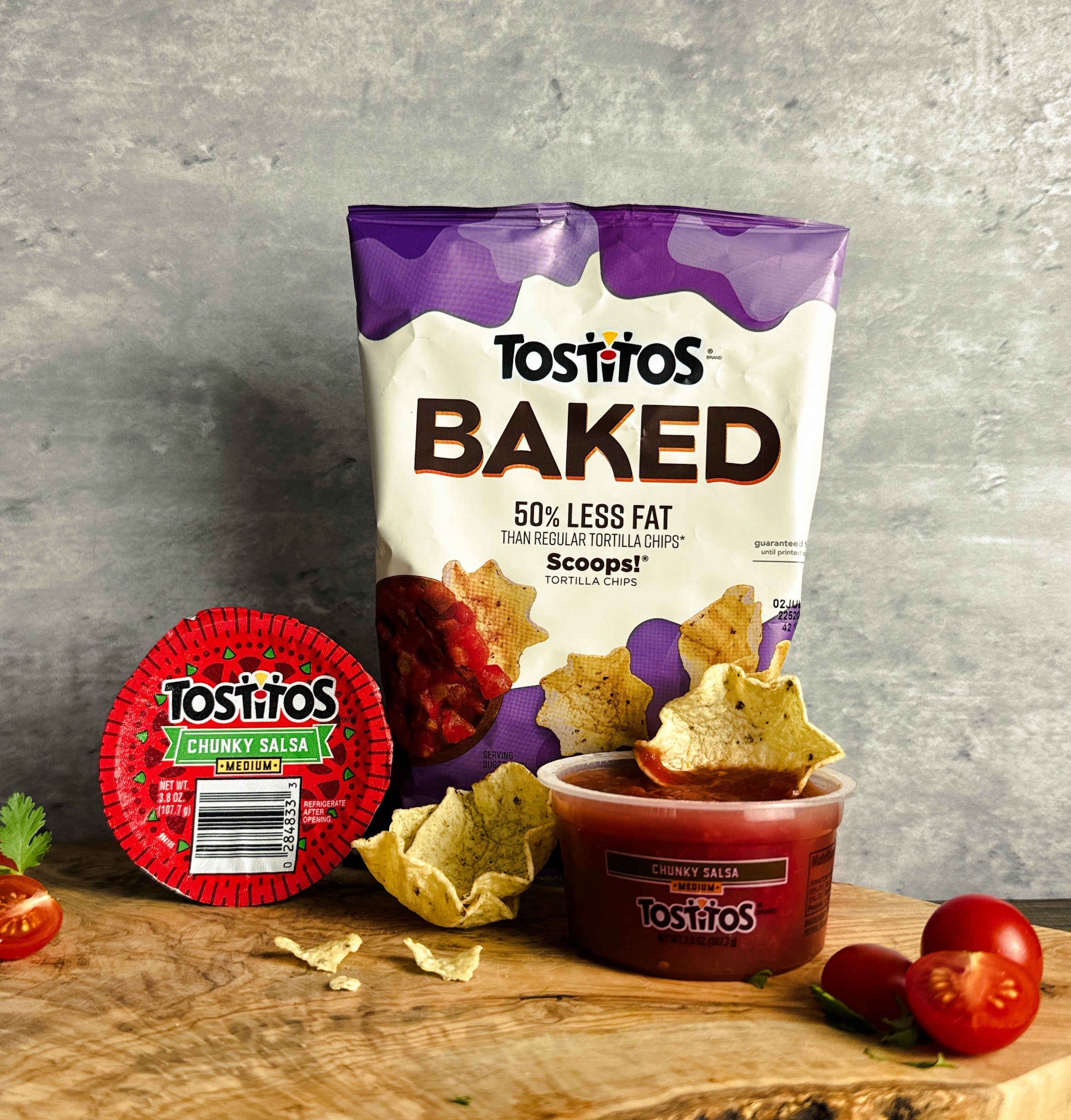 A pack of Tostitos Baked Scoop with Tostitos Chunky Salsa rests on a wooden surface alongside several tortilla chips and two sliced cherry tomatoes, promising a delightful snacking experience.