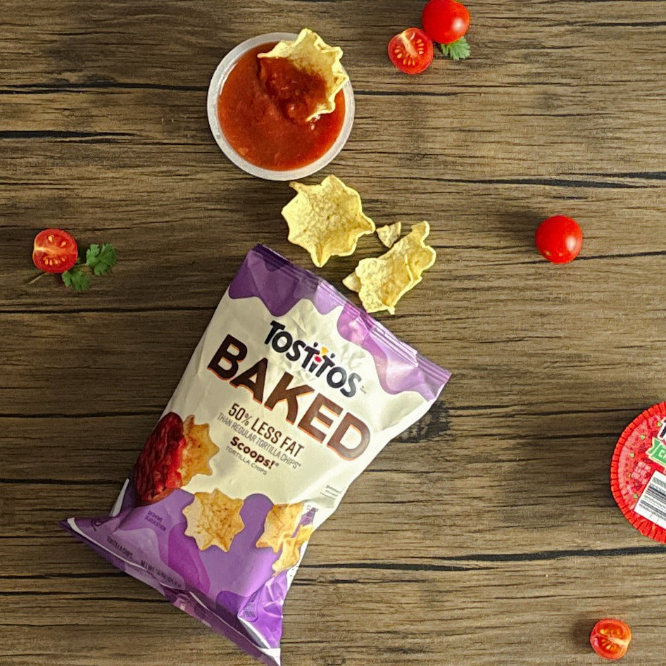 A bag of Tostitos Baked Scoop with Tostitos Chunky Salsa lies on a wooden surface, with some chips spilling out next to a container of Chunky Mild Salsa and cherry tomatoes, promising an enjoyable snacking experience.