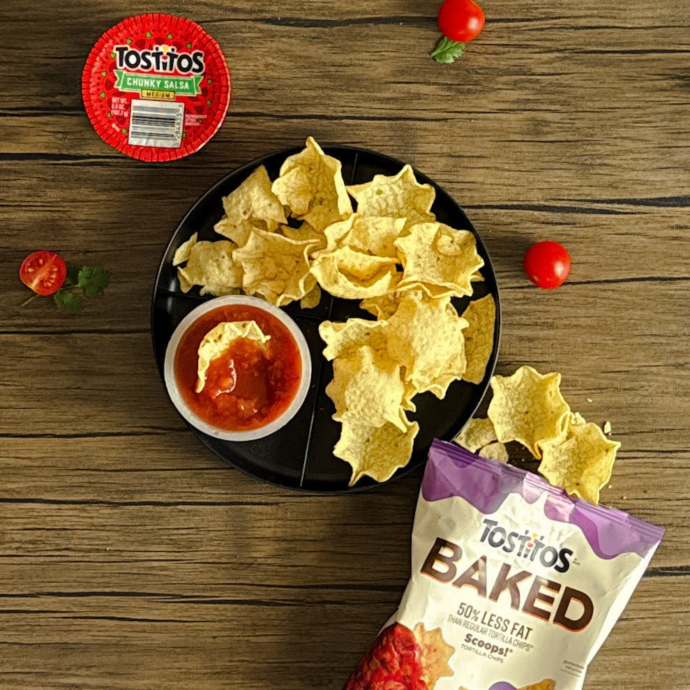 A plate of tortilla chips sits next to an open container of Tostitos Chunky Salsa. A bag of Tostitos Baked Scoops and some cherry tomatoes rest on a wooden surface, inviting you to elevate your snacking experience.