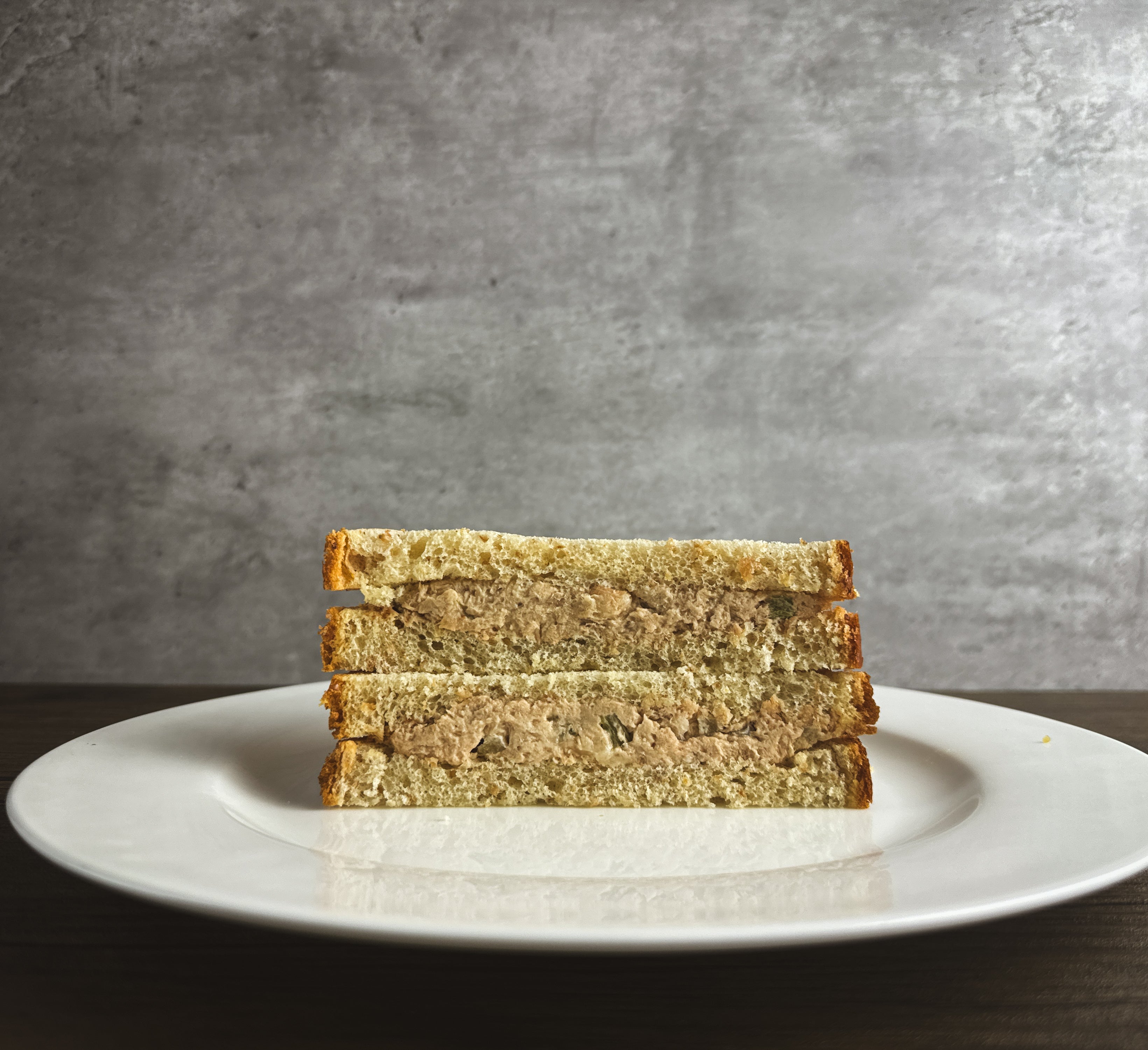 Stacked triangular Tuna Salad Sandwiches by Taylor Farms, made with yellowfin tuna and canola mayonnaise on white bread, served on a white plate against a gray background.