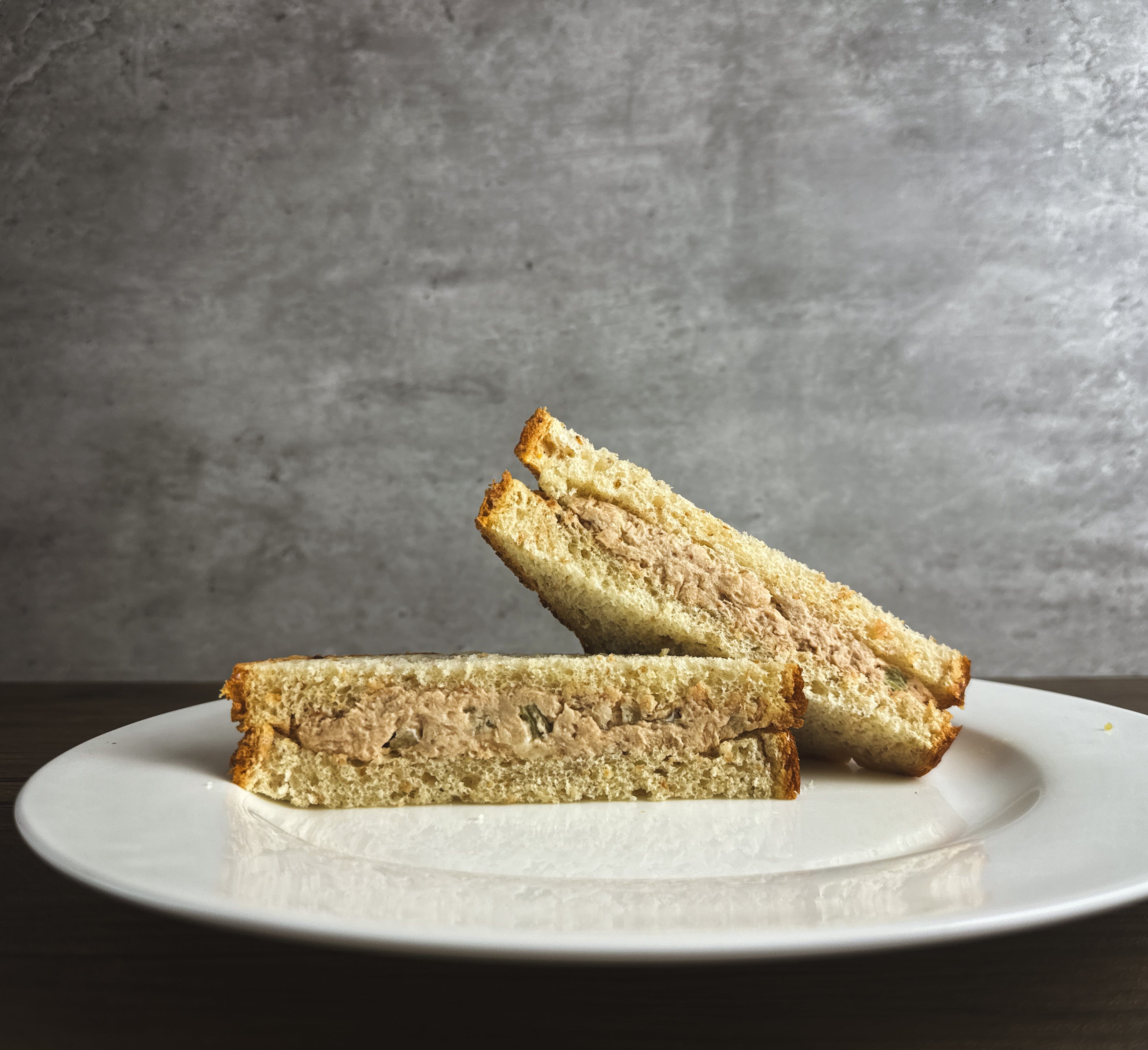 A Taylor Farms Tuna Salad Sandwich, divided into two triangular halves and placed on a white plate, rests on a wooden table with a gray concrete background.