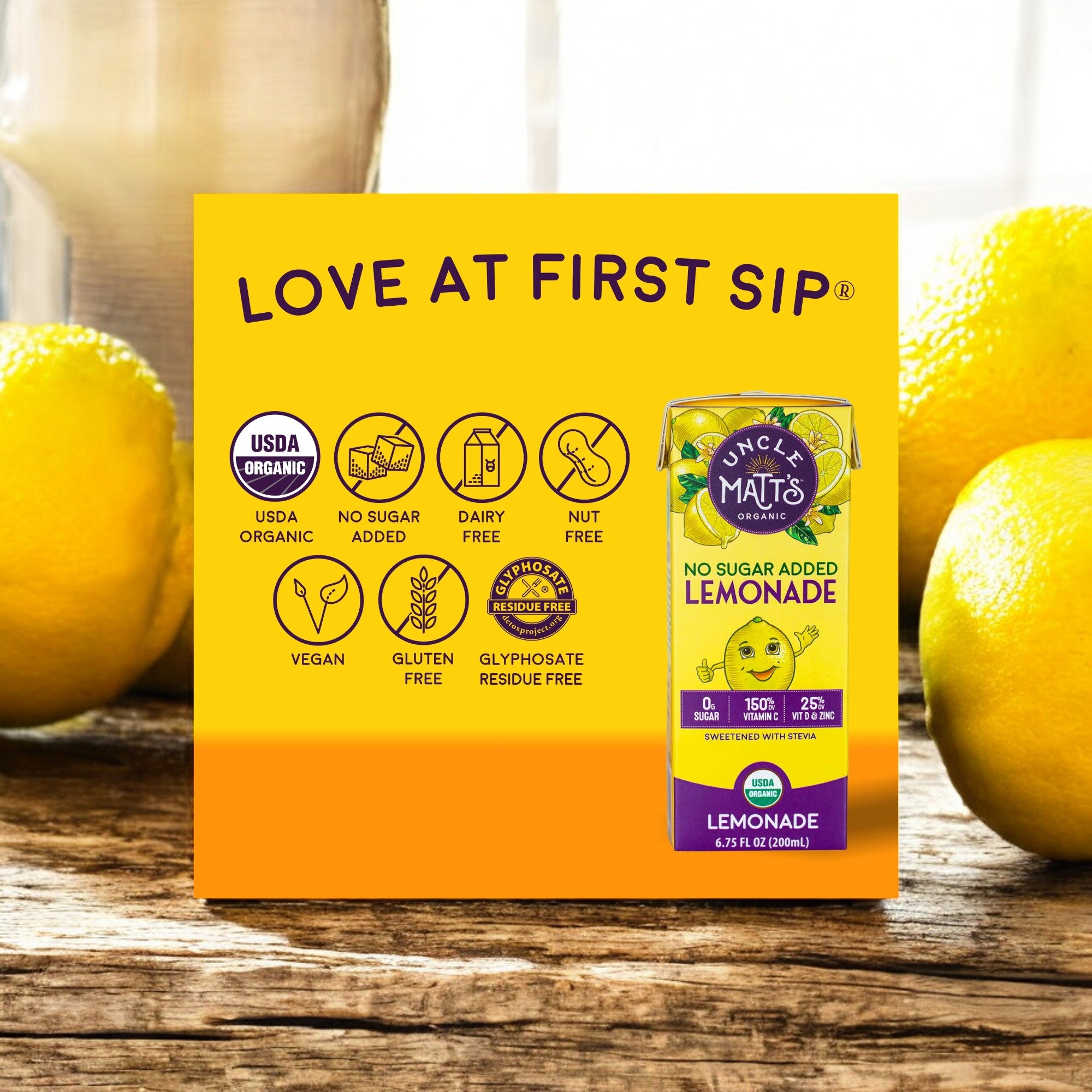 A box of "Uncle Matt's Lemonade Juice Box, 6.75 oz - 1 Count" sits on a wooden table with fresh organic lemons in the background. The box showcases features like "No Sugar Added," "USDA Organic," "Non-GMO," and various dietary logos.