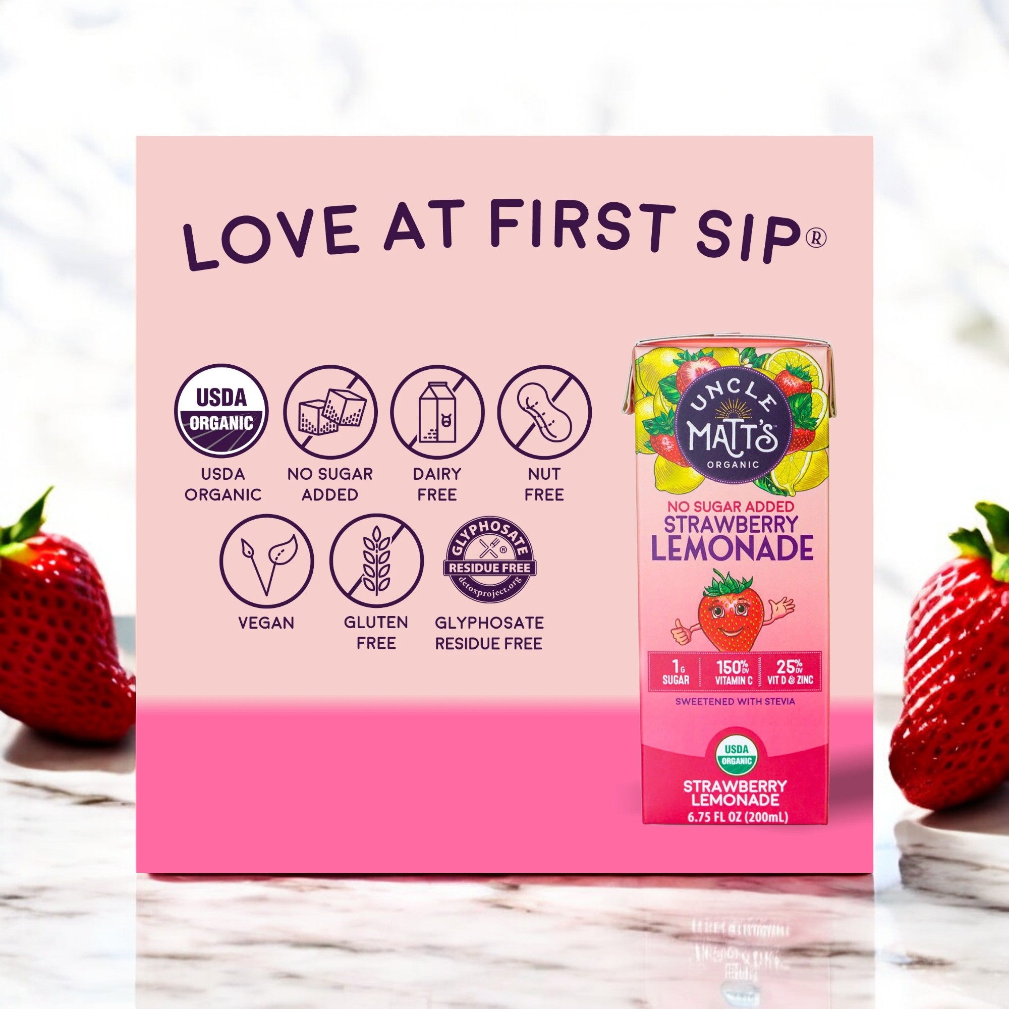 A carton of Uncle Matt's Strawberry Lemonade Juice Boxes - No Sugar Added, 6.75 oz is placed in front of a pink backdrop adorned with health icons and the text "LOVE AT FIRST SIP". Fresh strawberries are positioned on either side.