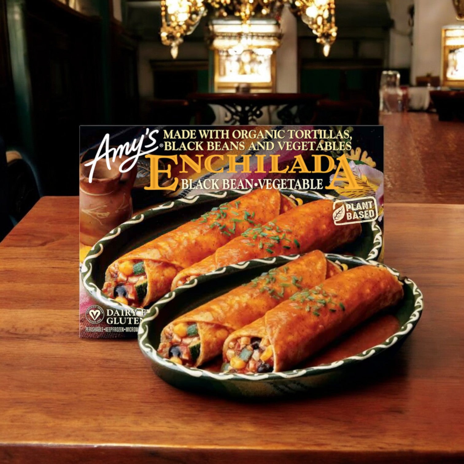 A package of Amy's Kitchen Enchilada with Spanish Rice & Beans Meal, 10 oz., is displayed on a wooden table in a dining room with a chandelier. The back of the package shows nutrition facts, ingredients, and preparation instructions.