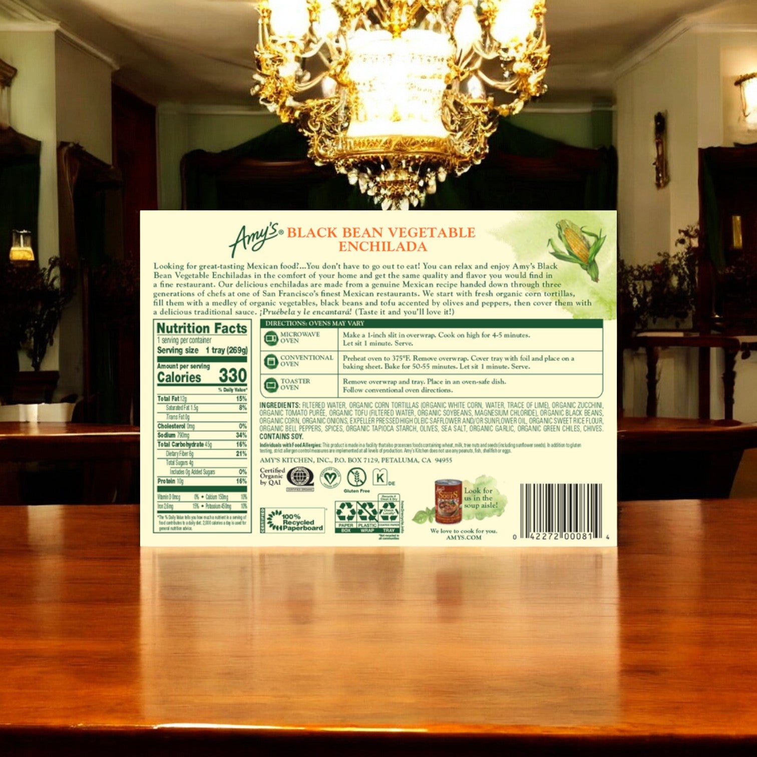 A package of Amy's Kitchen Enchilada with Spanish Rice & Beans Meal, 10 oz., is displayed on a wooden table in a dining room with a chandelier. The back of the package shows nutrition facts, ingredients, and preparation instructions.