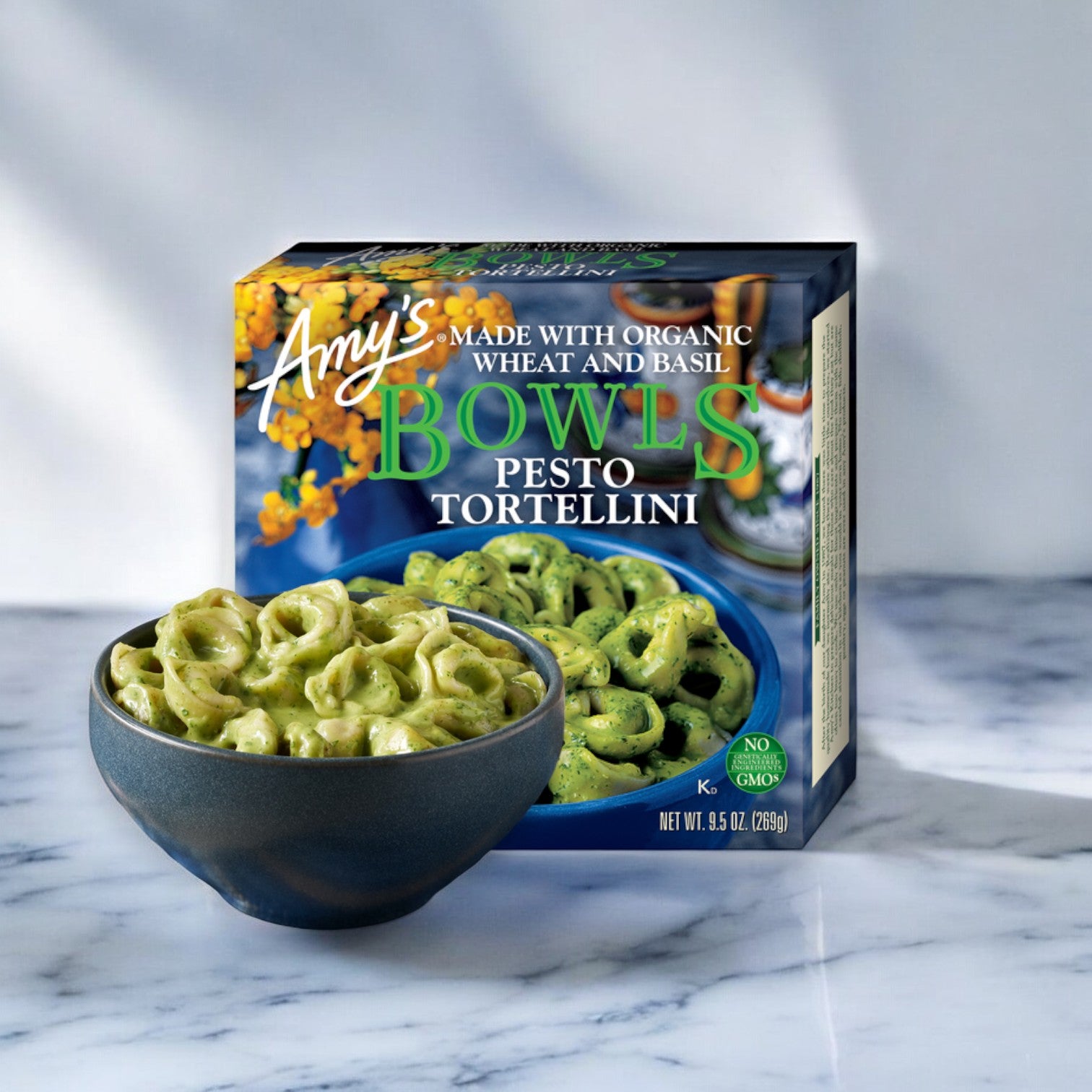 Box of Amy's Kitchen Bowls Pesto Tortellini, 9.5 oz. - 1 Count with a filled bowl in front, placed on a marble surface. An Italian-inspired meal made with organic ingredients by Amy's.