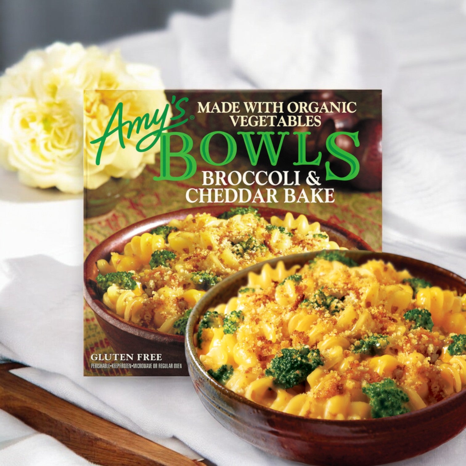 A frozen meal package labeled "Amy's Kitchen Broccoli & Cheddar Bake Meal Bowl, 9.5 oz. - 1 Count," showing a bowl of pasta with organic black beans, broccoli, and cheddar, placed on a wooden surface with white flowers in the background.