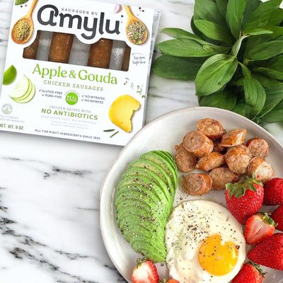 A plate with sliced avocado, a sunny-side up egg, strawberries, and fully cooked Amylu Apple & Gouda Chicken Sausage made from antibiotic-free chicken sits beside its box on a marble surface.