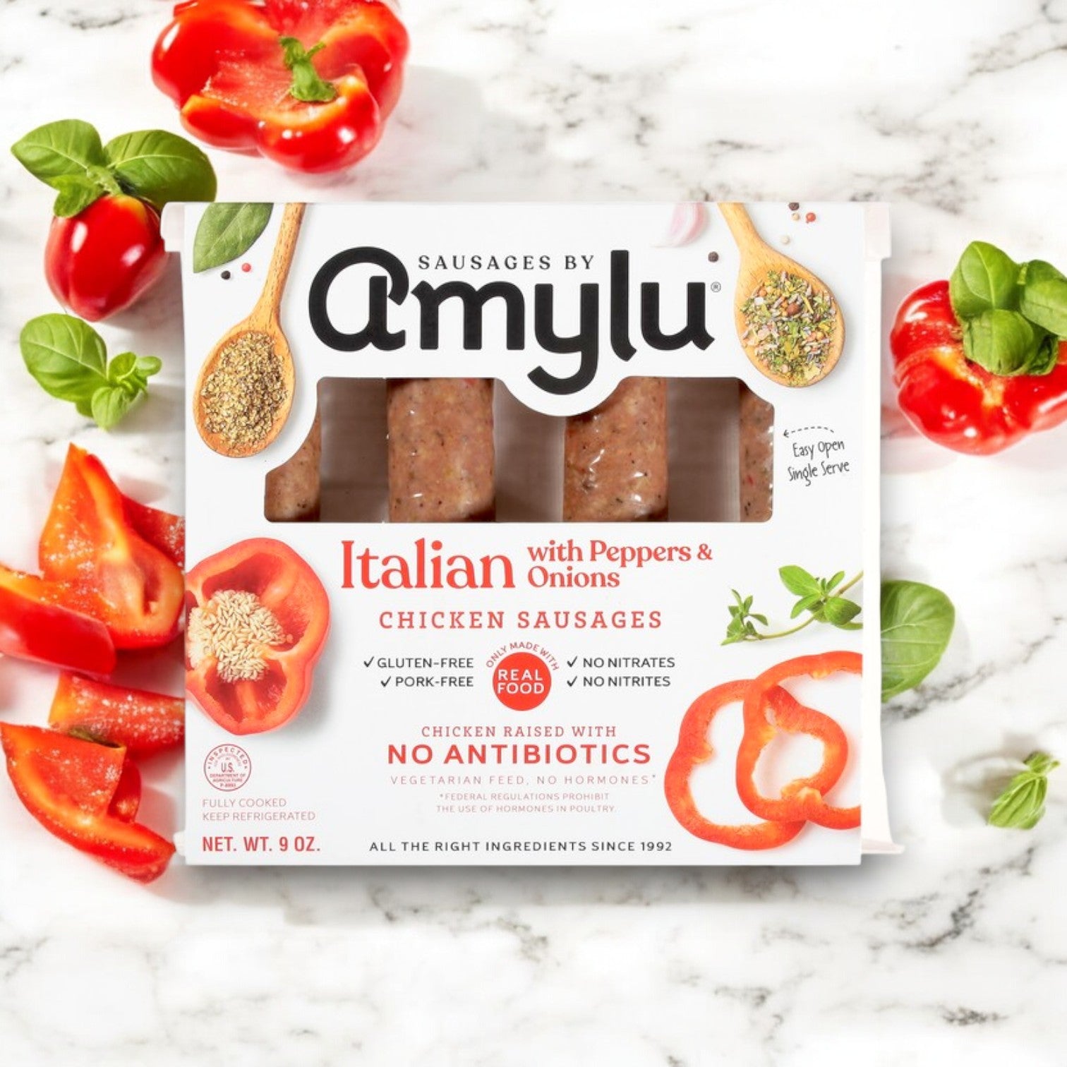 A package of Easy Lunches Amylu Italian Chicken Sausage with Peppers & Onions – 3 Packs is shown, surrounded by sliced red bell peppers and cherry tomatoes on a white marble surface.