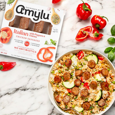 A package of Amylu Italian Chicken Sausage with Peppers & Onions – 9 oz is next to a bowl of rice mixed with sliced sausage, cherry tomatoes, and zucchini on a marble surface.