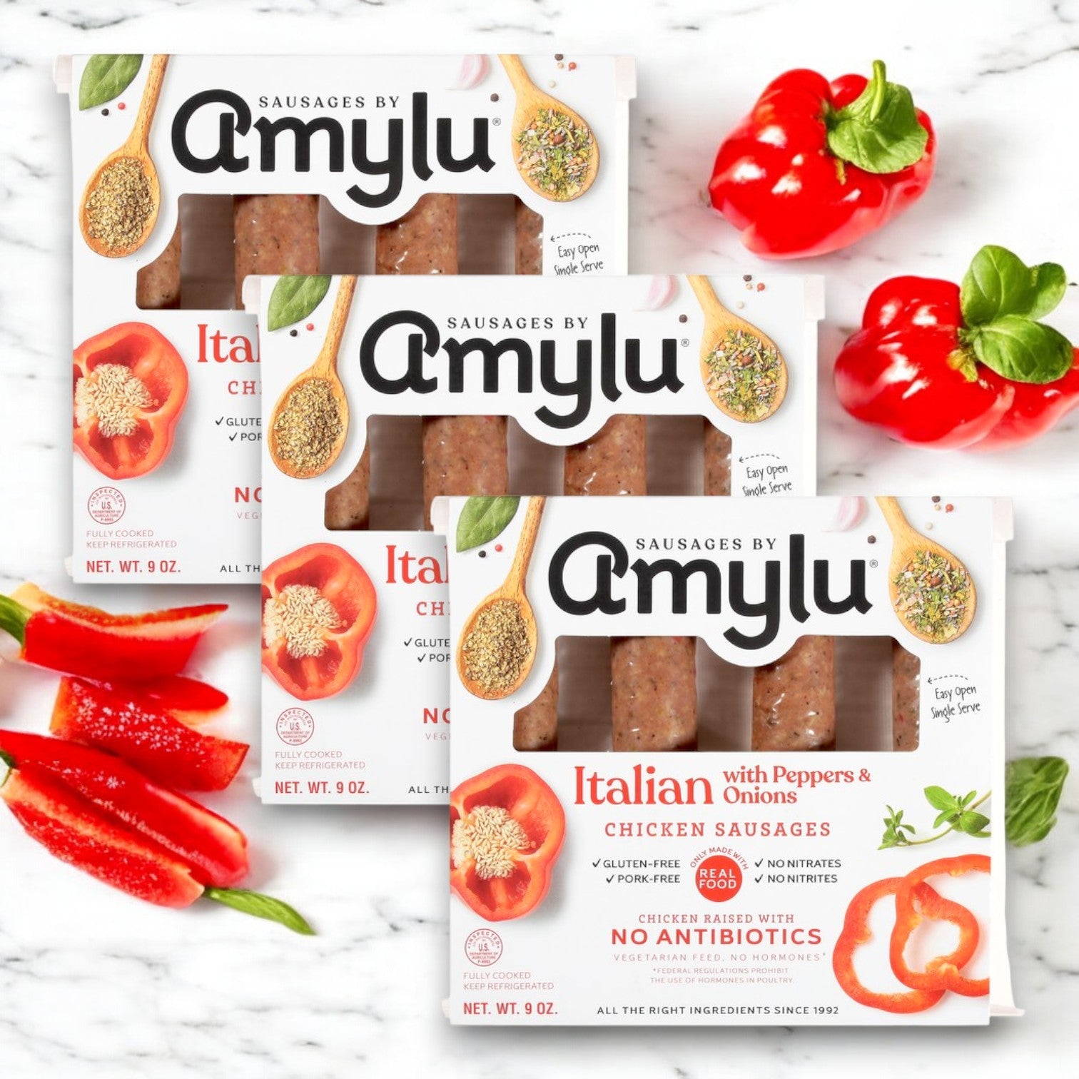 Three packs of Easy Lunches Amylu Italian Chicken Sausage with Peppers & Onions are arranged on a marble surface, accented by fresh red bell peppers and basil leaves.