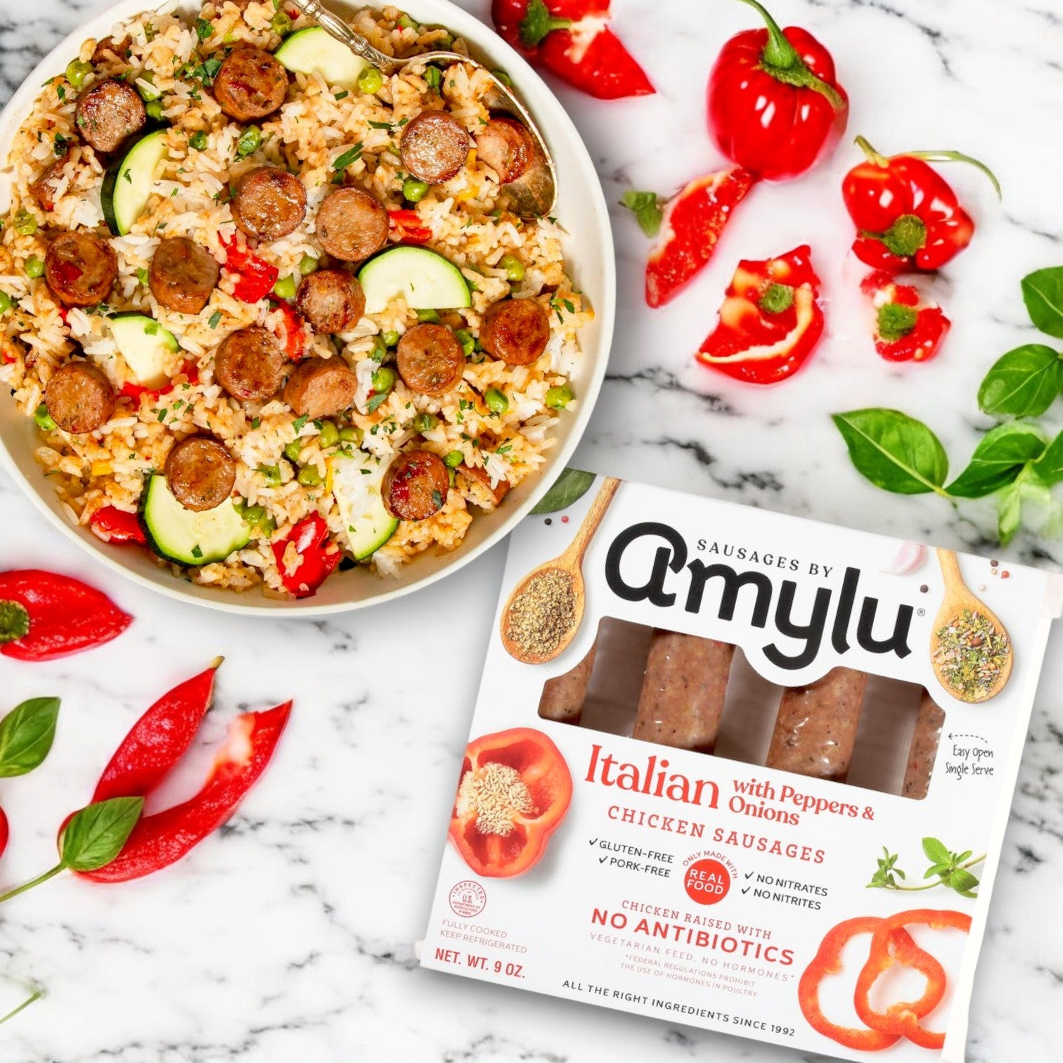 A bowl of rice with sliced Amylu Italian Chicken Sausage with Peppers & Onions by Easy Lunches sits on marble beside its 3-pack package, with chopped red peppers, onions, zucchini, and fresh herbs.