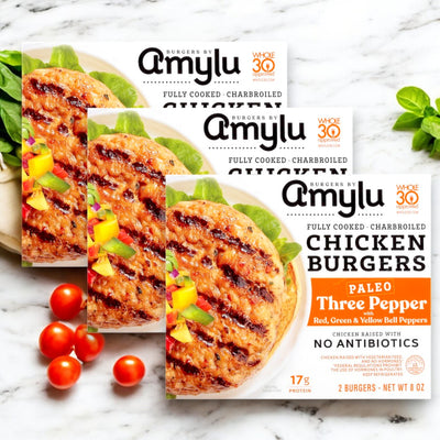 Three boxes of Amylu Paleo Charbroiled Three Pepper Chicken Burgers, featuring vibrant burger and bell pepper images on the packaging, surrounded by fresh tomatoes and basil. Made with antibiotic-free chicken, these delicious 24 oz. patties are a perfect gluten-free meal choice.