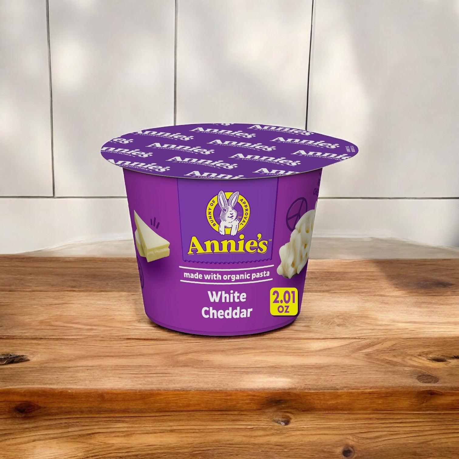 A 2.1-ounce cup of Annie's White Cheddar Mac and Cheese Cups, made with organic pasta, featuring a purple lid and container, sits on a wooden surface against a blurred background. This ready-to-microwave mac and cheese from Annie's is the perfect option for a quick and delicious meal.