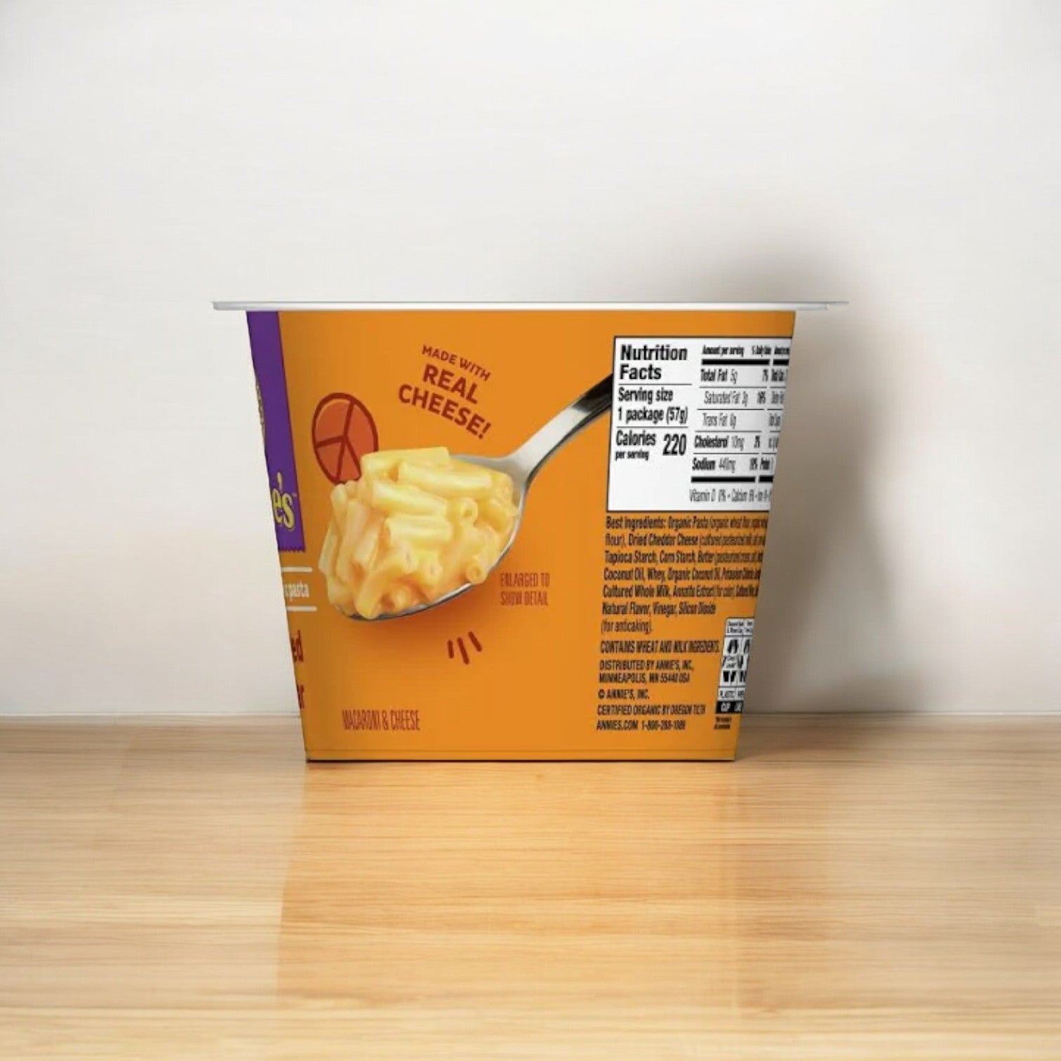 An Annie's Real Aged Cheddar Microwave Mac and Cheese Cup, 2.01 oz - 1 Count, featuring a "Made with Real Cheese!" label and displaying nutritional information on its side, placed on a wooden surface against a plain wall.