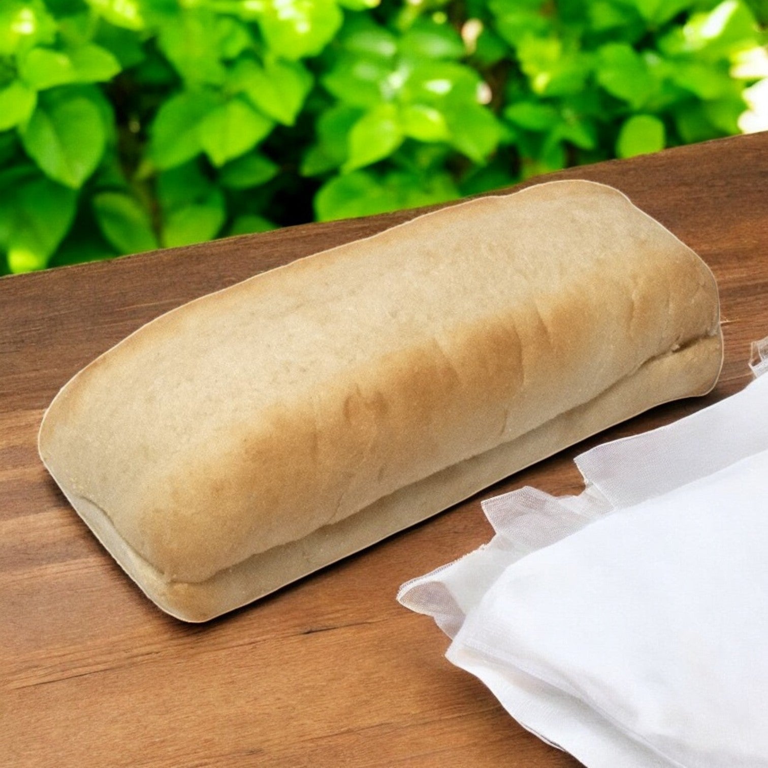 A sealed pack of Easy Lunches Italian Beef Hoagie Buns sits on a wooden table, surrounded by lush greenery. A white cloth peeks out beside it, evoking the warmth and tradition of hearth-baked goods.