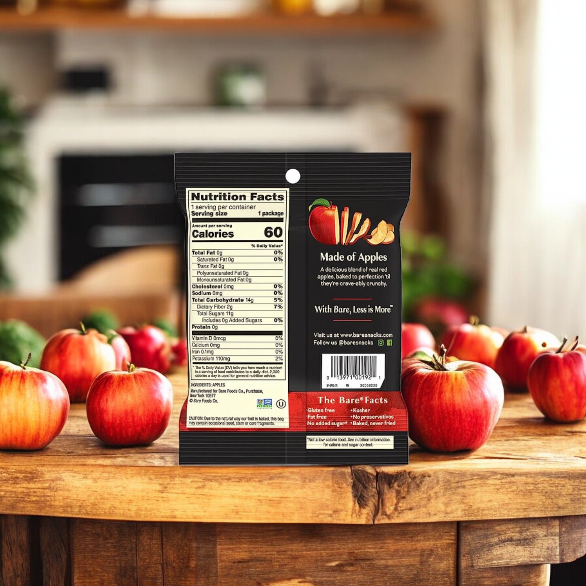 A packet labeled "Bare Baked Crunchy Crispy Red - Apple Flavor, 0.53 oz., 1 Count" from Easy Lunches, with visible nutrition facts and ingredients, sits on a wooden table with several fresh apples in the foreground and a kitchen setting in the background, showcasing this healthy snack option.