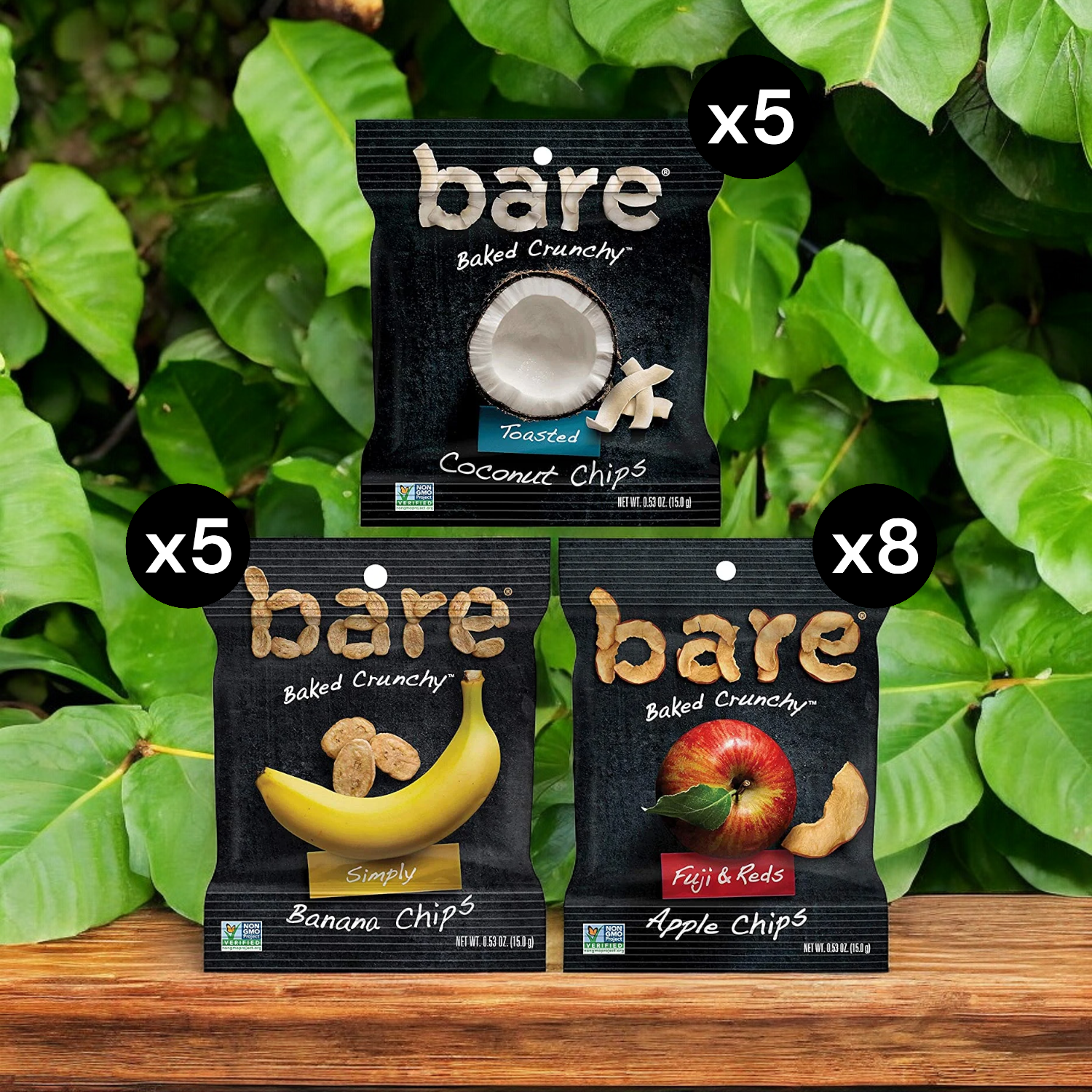 Three types of Easy Lunches Bare Baked Crunchy Variety Pack Chips (0.53 oz., 18 pk.) on a wooden surface: 5 bags of Toasted Coconut Chips, 5 bags of Banana Chips, and 8 bags of Apple Chips; green leaves in the background. Enjoy this healthy snack variety pack with flavorful fruit chips perfect for any time.