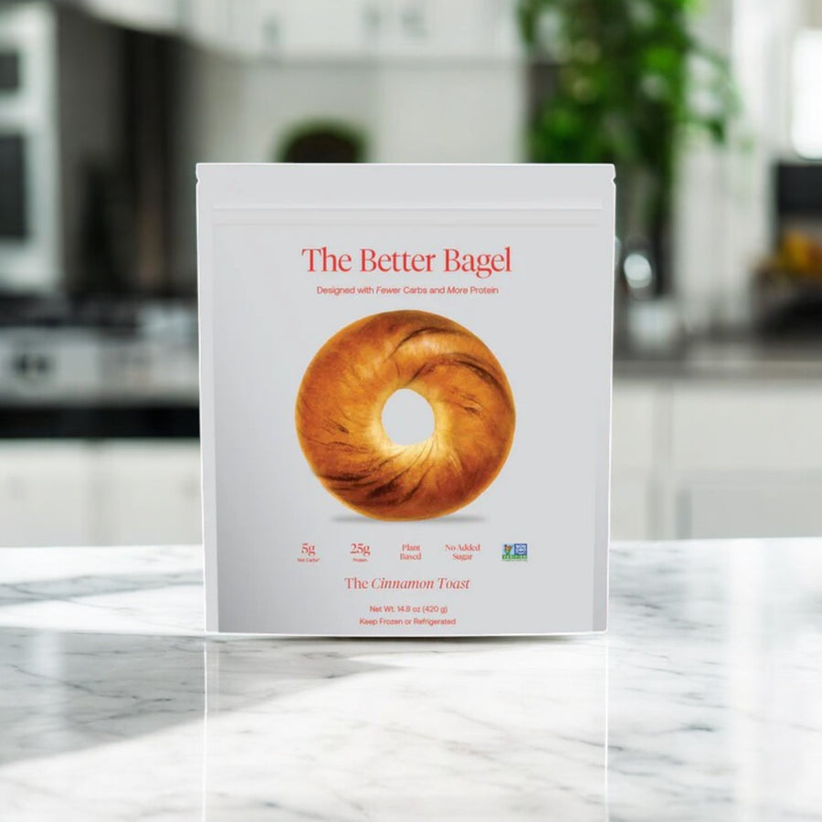 A white bag labeled "Better Brand – The Cinnamon Better Bagel" (1 bag, 4 total bagels) with Low-Carb and High-Protein highlights is shown on a kitchen counter, background blurred. In stock and ready to ship from BetterBrand!.