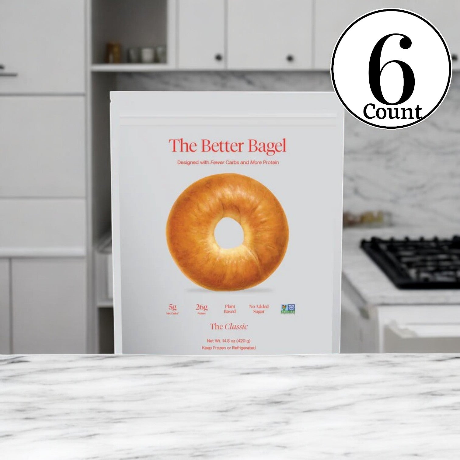 A package labeled "BetterBrand - The Classic Better Bagel" rests on a marble countertop in a kitchen, featuring its classic bagel flavor with details of being high protein, low carb, and containing no added sugar, all within the 6-bag pack totaling 24 bagels.