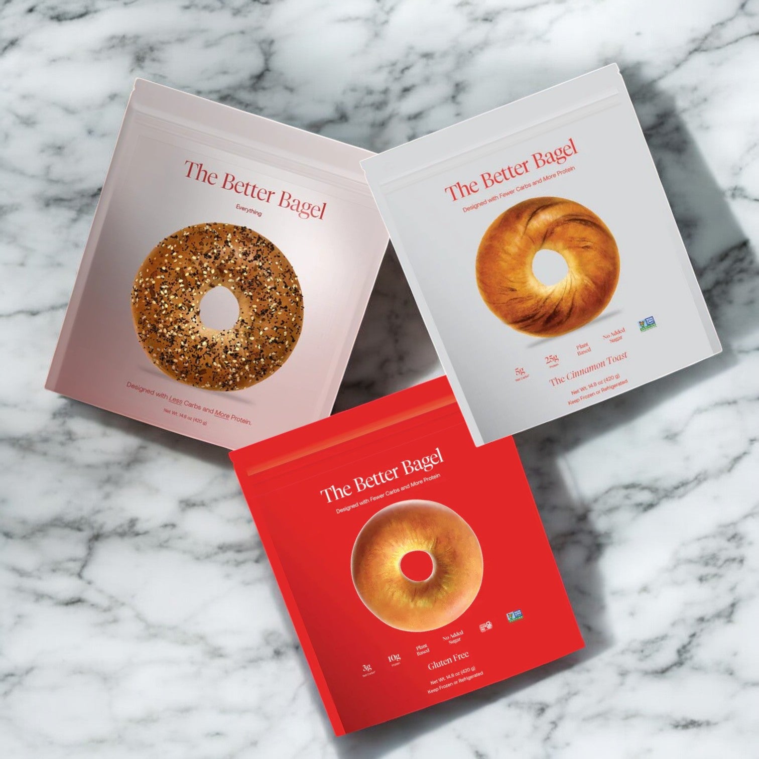 Displayed on a marble surface, the BetterBrand - The BetterBrand 3 Pack Combo features three packages, each highlighting a distinct bagel variety: everything, cinnamon toast, and plain—all made with premium ingredients by BetterBrand.