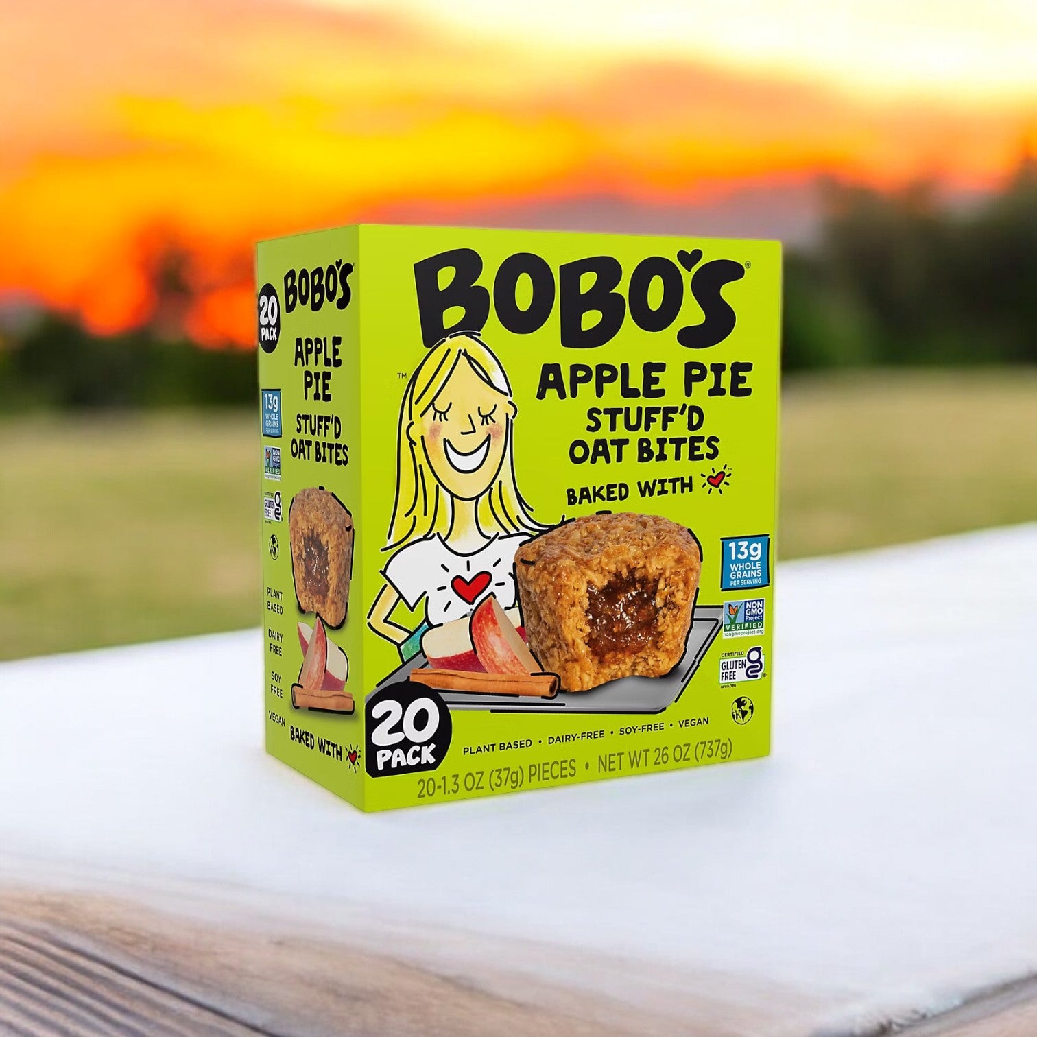 A box of Bobo's Apple Pie Filled Bites (20 count) sits on a table, set against a vibrant sunset. Ideal for an on-the-go snack, the plant-based packaging proudly highlights 13g of whole grain oats per serving.