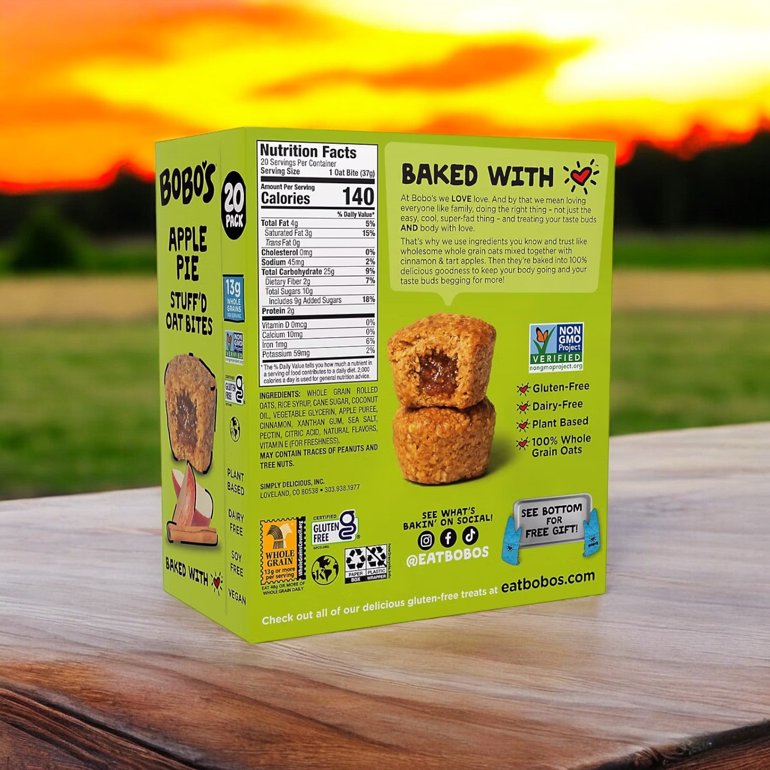 A box of Bobo's Apple Pie Filled Bites, containing 20 pieces, rests on a wooden surface against a sunset backdrop. These delicious bites are an ideal wholesome snack for those on the go, proudly showcasing labels that affirm they are gluten-free, plant-based, and non-GMO.