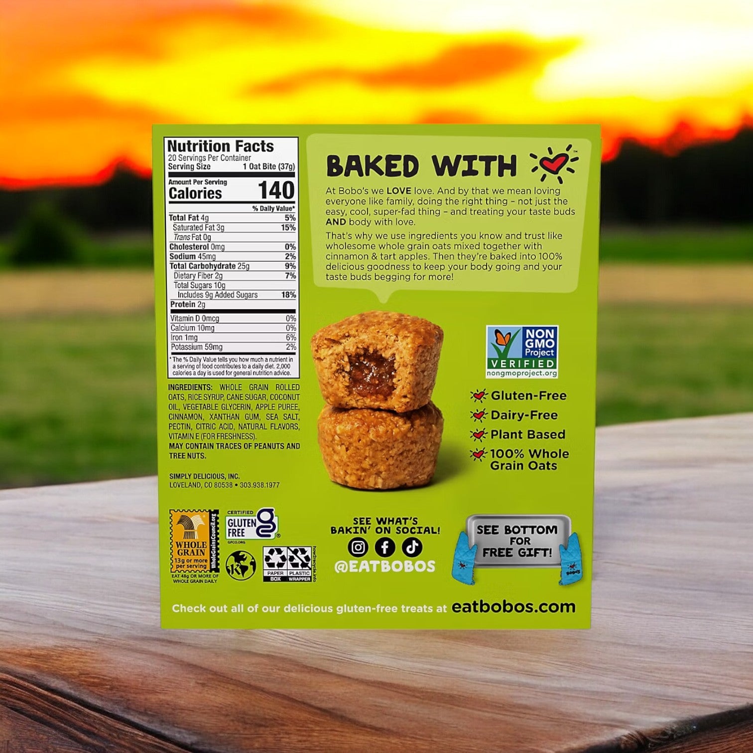 A box of Bobo's Apple Pie Filled Bites, 20 count, rests on a wooden table, highlighting its gluten-free nutrition facts and product details against a sunset backdrop. Ideal as a wholesome snack for those on the move, these bites provide convenience without sacrificing quality ingredients.