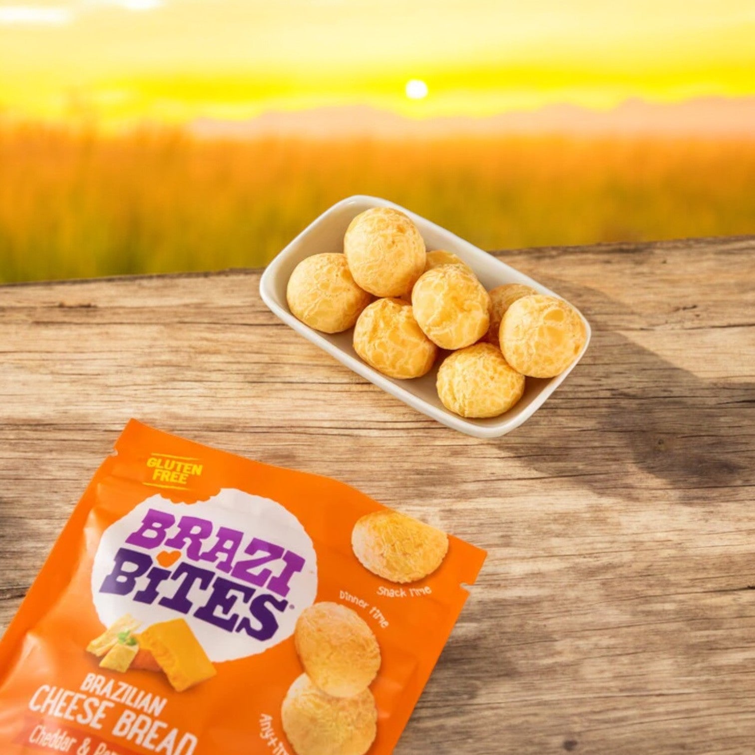 A bowl of Brazilian Cheese Bread in a round shape is placed on a wooden surface alongside an orange package of Brazi Bites Cheddar & Parmesan, showcasing the savory flavors. In the background, a sunset over grassy fields enhances the allure of this gluten-free cheese bread from Brazi Bites.