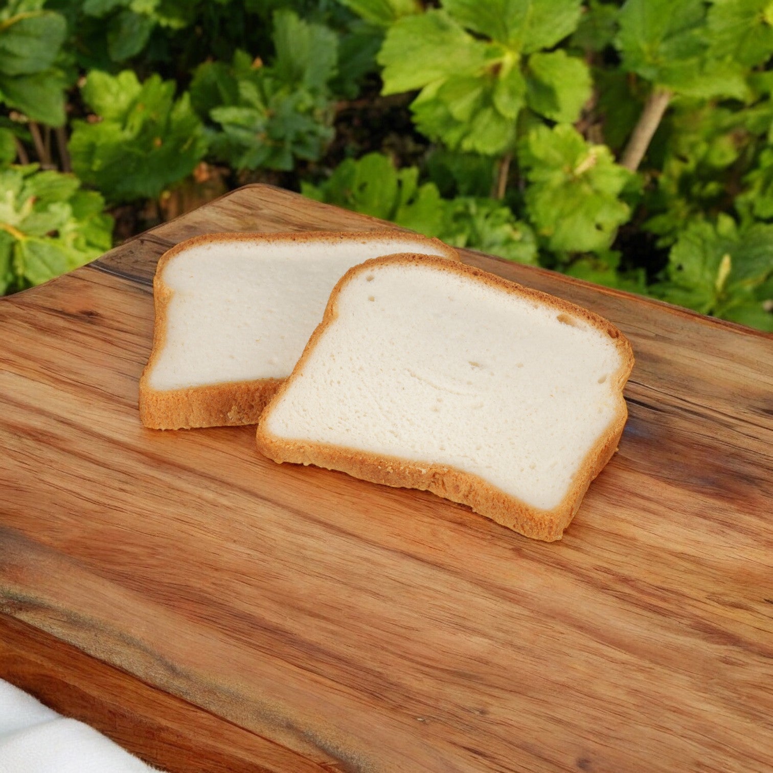 Two slices of Udi's Bread White Soft Sandwich Gluten Free rest on a wooden board, surrounded by lush green foliage in the background.