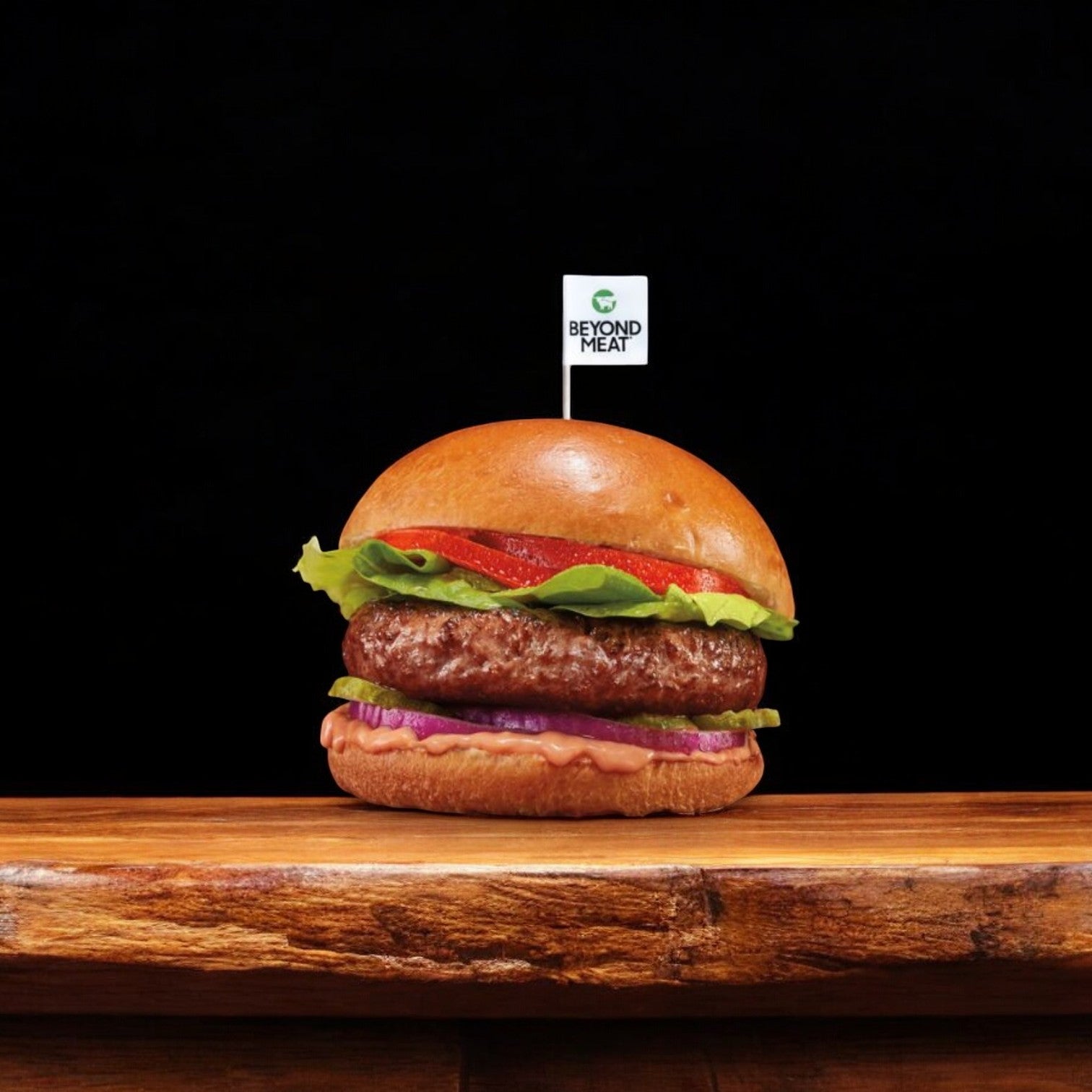 Introducing the Burger Vegetarian Beyond Patty from Beyond Meat: a delectable plant-based burger topped with fresh lettuce, ripe tomato, and zesty onion, all served on a traditional bun. This meat-free meal is crowned with a small "Beyond Meat" flag, making it an irresistible choice for those opting for plant-based alternatives. Displayed against a sleek black background, it's truly an appetizing sight.