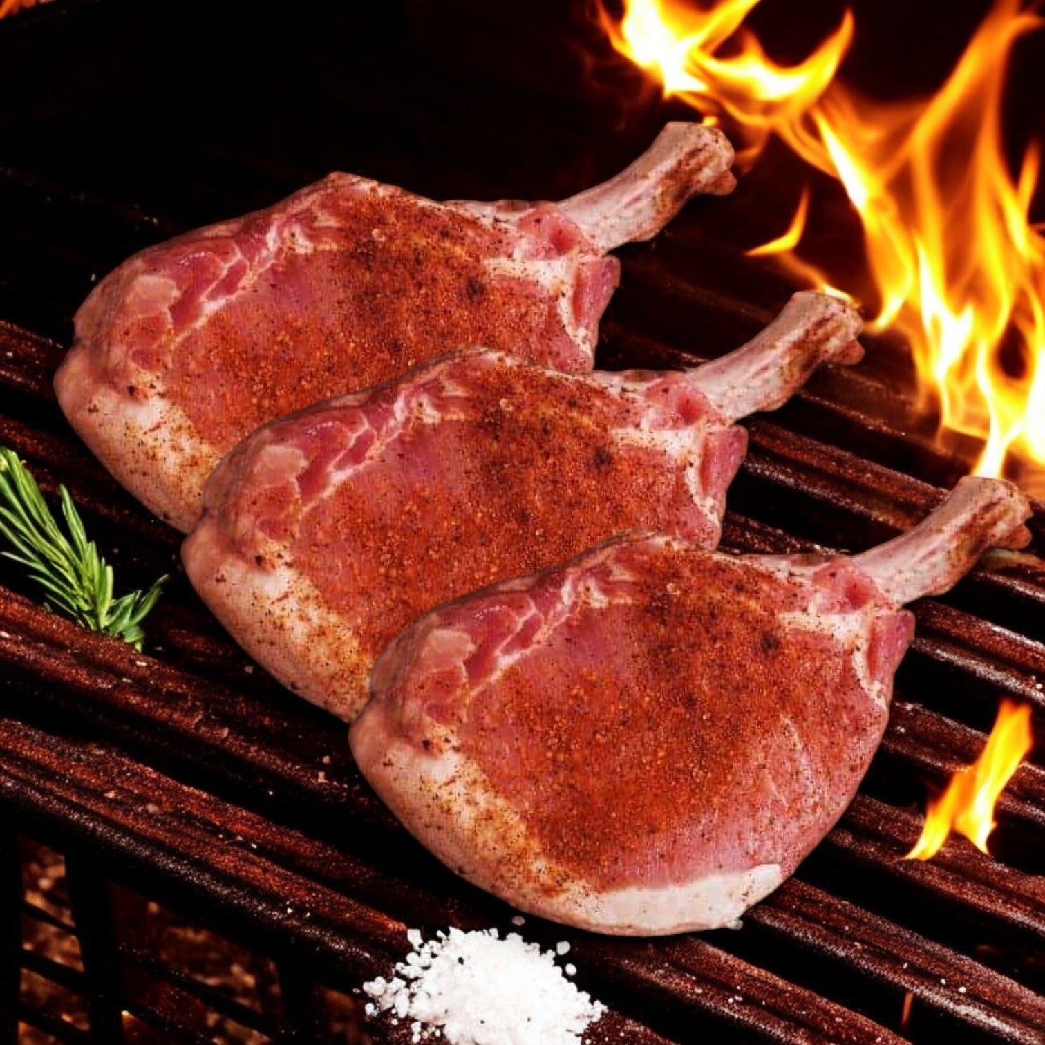 Three Mino Prime Steaks' 12 oz French Pork Chops sizzle on the grill with dancing flames. A sprig of rosemary and coarse salt stand ready to enhance their flavor, capturing Mino Prime Steaks' dedication to premium quality.