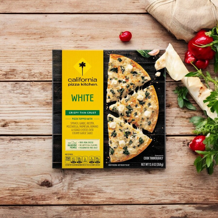 A package of California Pizza Kitchen White Thin Crust - 1 Count pizza from California Pizza Kitchen is displayed on a wooden surface, encircled by fresh ingredients such as tomatoes and herbs. The gourmet pizza is partially sliced, revealing its rich toppings of various cheeses and spinach.