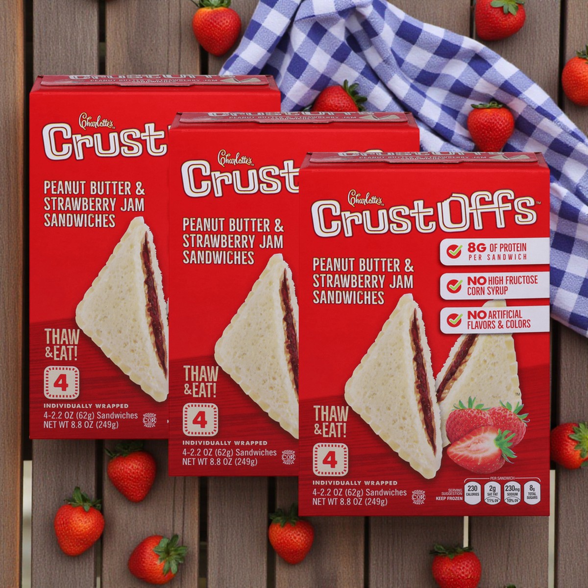 Three boxes of Crust Off's Charlotte’s Crust Offs Sandwiches – Peanut Butter & Strawberry Jam, 2.2 oz each, are displayed on a wooden surface with fresh strawberries and a blue checked cloth—ideal for quick, tasty lunchbox snacks.