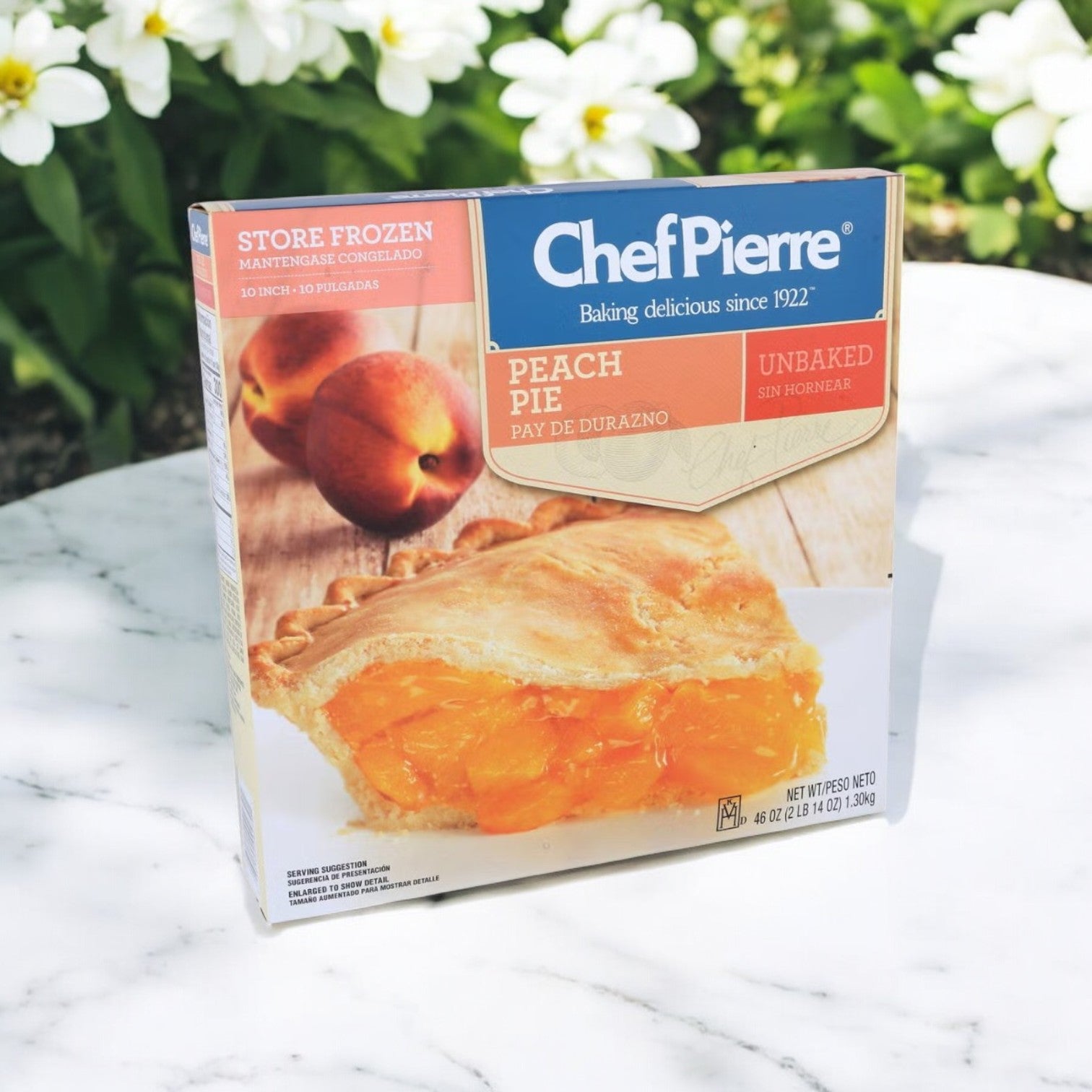 A 46 oz. box of Chef Pierre Peach Pie, showcasing a peach slice and whole peaches, rests on marble. Adorned with white flowers and greenery, the package promises a fresh and delightful frozen unbaked dessert experience.